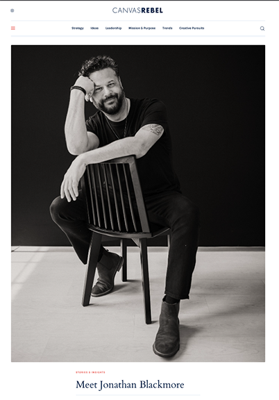 A man with a beard and curly hair sitting on a wooden chair, smiling, with his left arm resting on the back of the chair and his right arm on his knee, wearing a black t-shirt, black pants, and boots, in front of a black background.