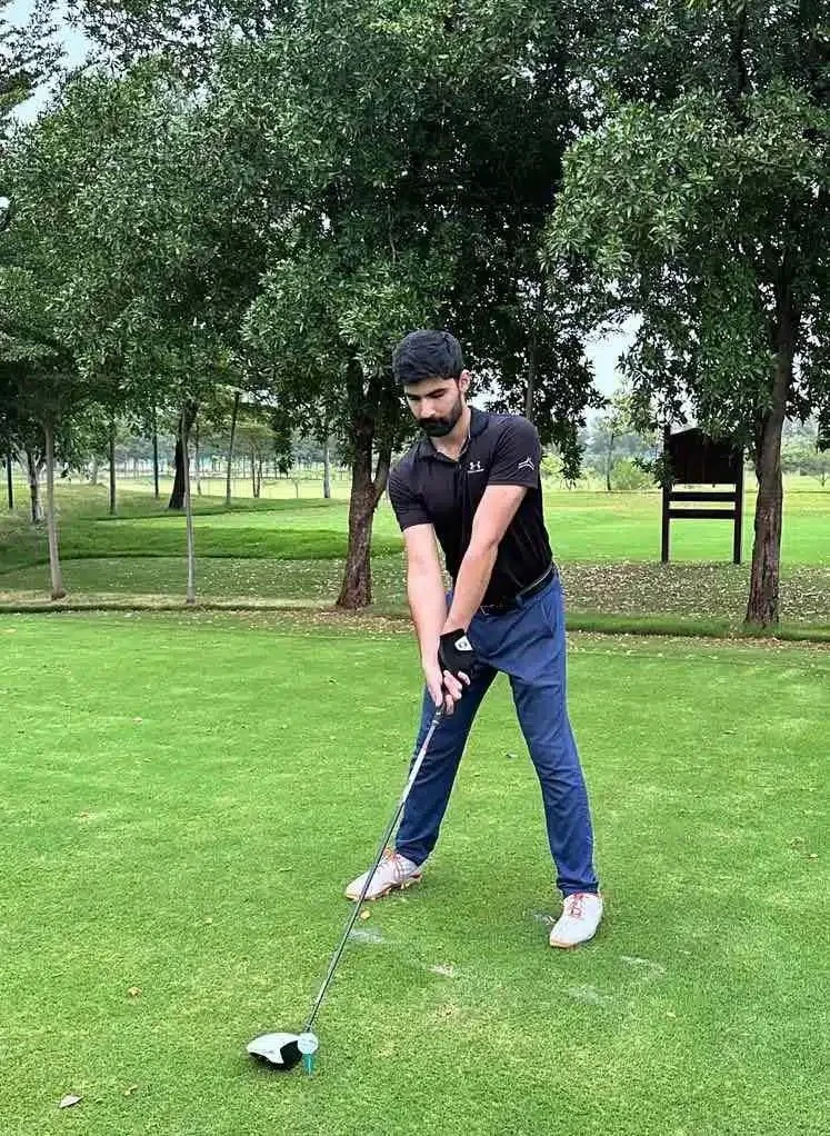 A man with a beard and dark hair, wearing a black polo shirt, blue pants, and white shoes, is preparing to tee off on a golf course. He is holding a golf club, focusing on the ball, with trees and a golf course in the background.