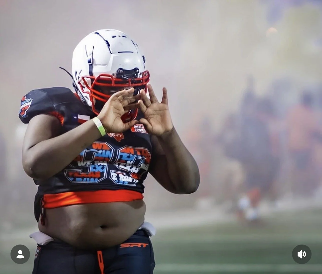 A young football player wearing a helmet and a jersey, making a gesture with his hands on a football field with smoke or fog in the background.