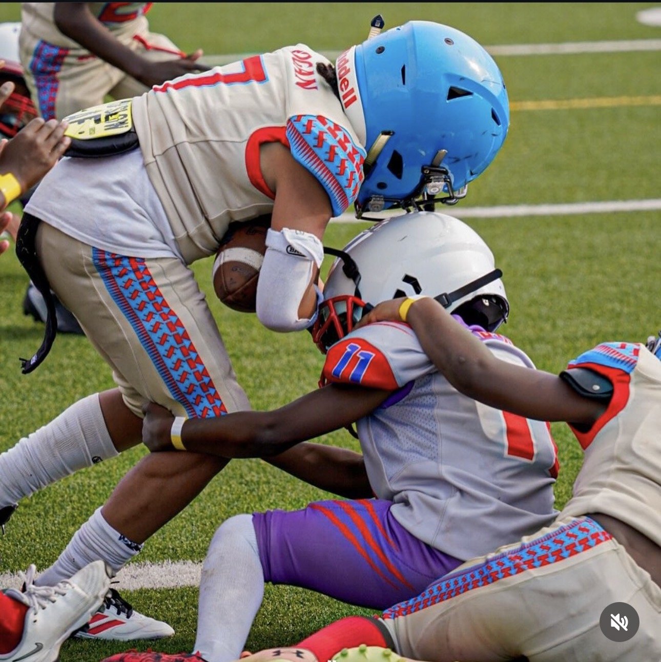 Two American football players in uniform during a game, one standing and reaching forward, the other on the ground, holding a football, on a grassy field.