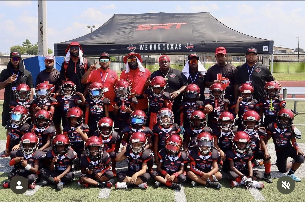 Youth football team and coaches posing on a football field under a tent that says "We Run Texas" and "SRT" with players in black and red uniforms, helmets, and sunglasses.