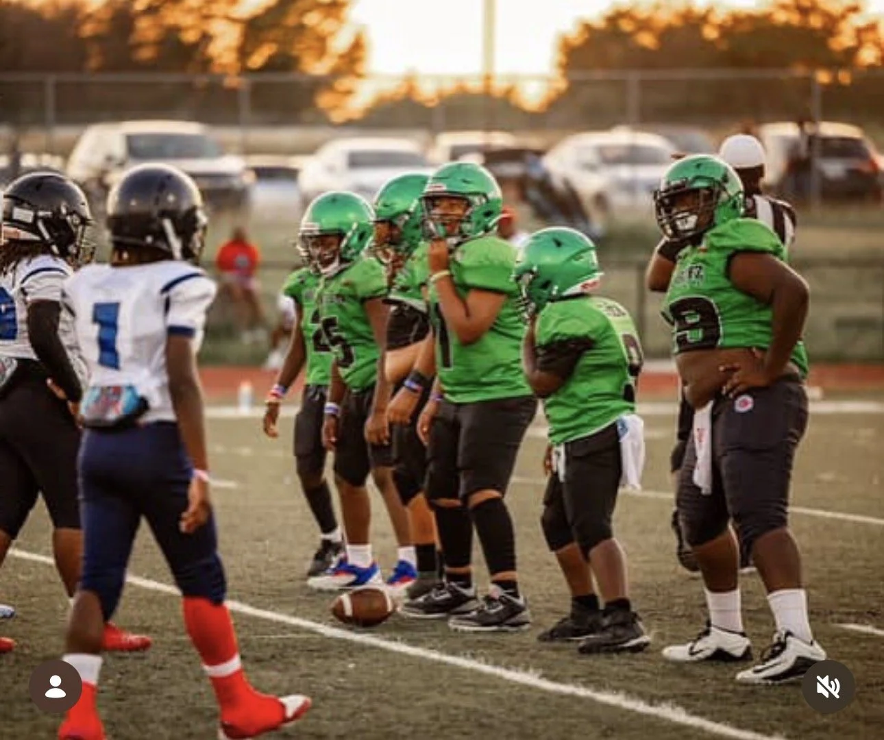 Youth football game on a field at sunset with players in green and black, and white and blue uniforms, standing at the line of scrimmage.