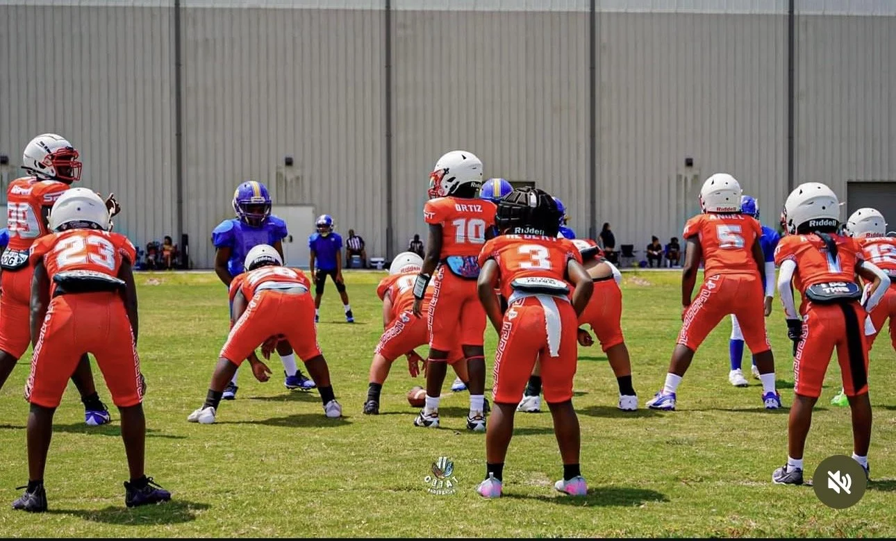A youth football team in red uniforms practicing on a grass field with other players and officials in the background.