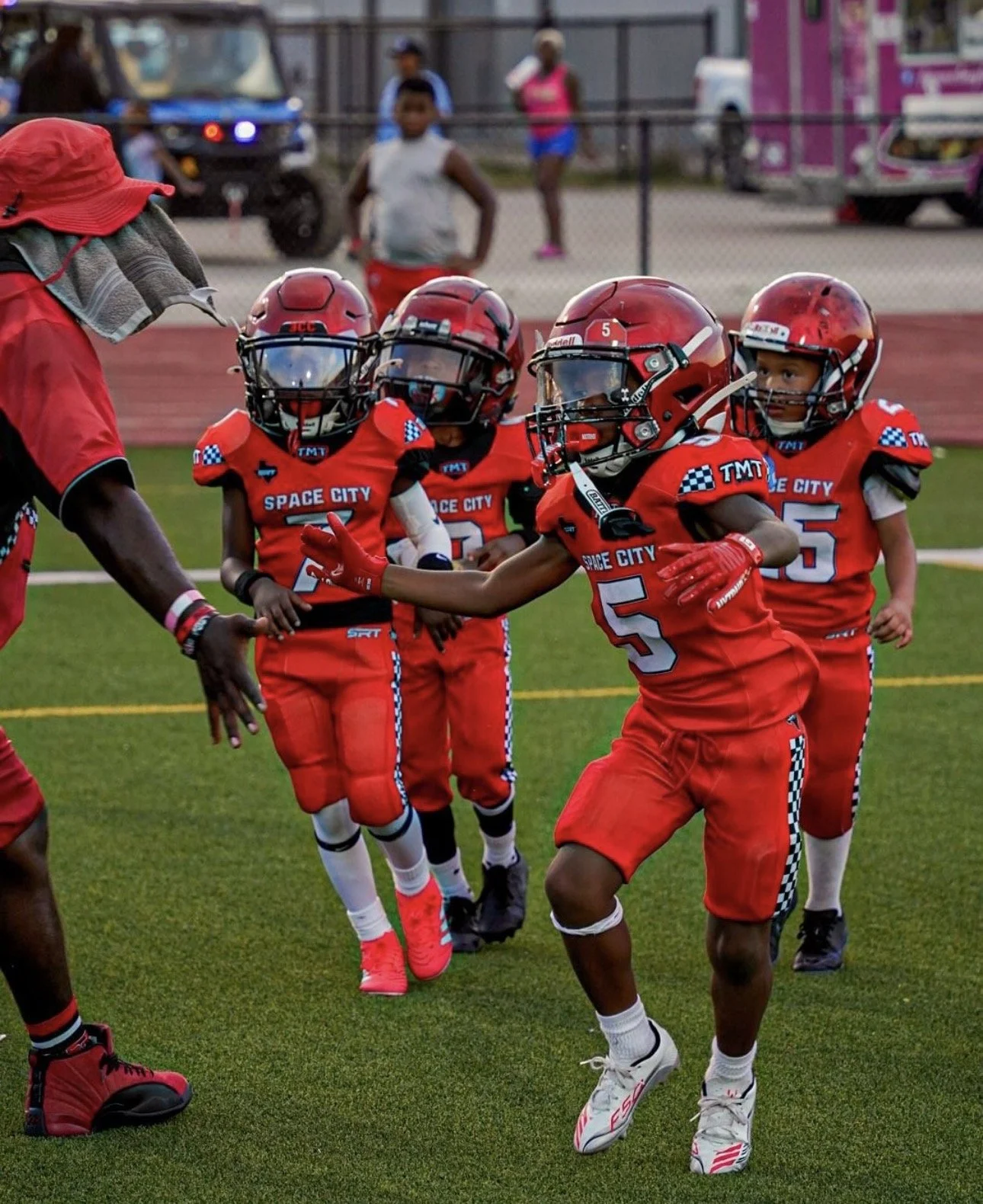 Young boys in red football uniforms and helmets practicing with coach on a grass field.