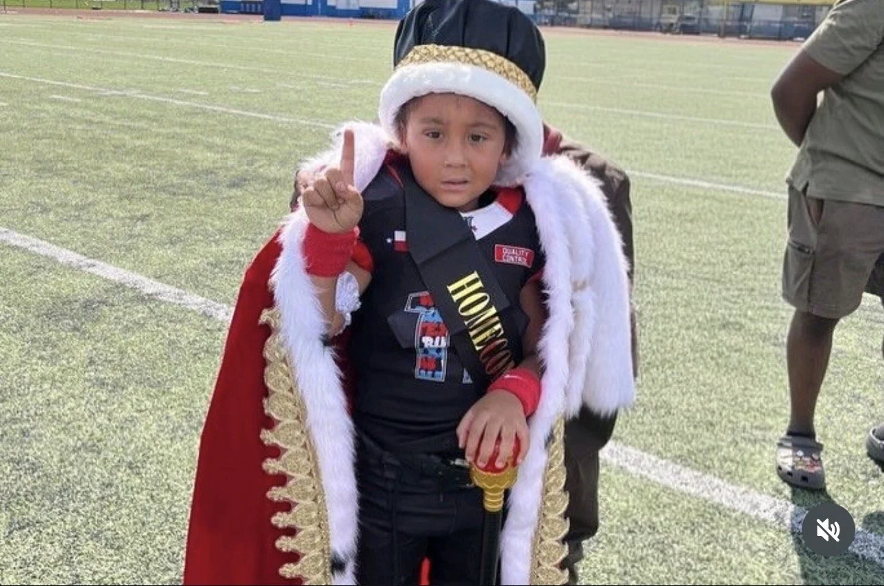 A young boy dressed as a king with a crown, red cape, and a sash reading 'Honorable' stands on a sports field, making a gesture with his finger. He appears surprised or confused.