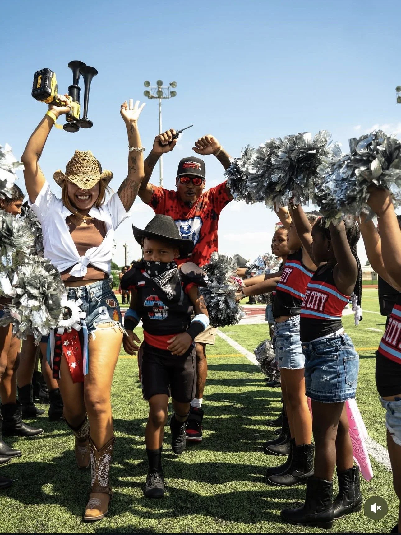 Cheerleaders and children participating in a cheerleading routine on a sports field during a sunny day, with spectators and a light pole in the background.