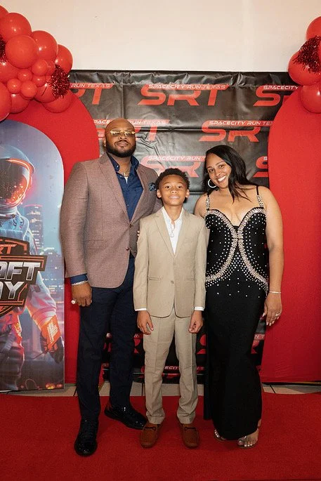 A man, a woman, and a young boy standing together at a red carpet event with a SpaceX and Starcraft-themed backdrop and red balloon decorations.