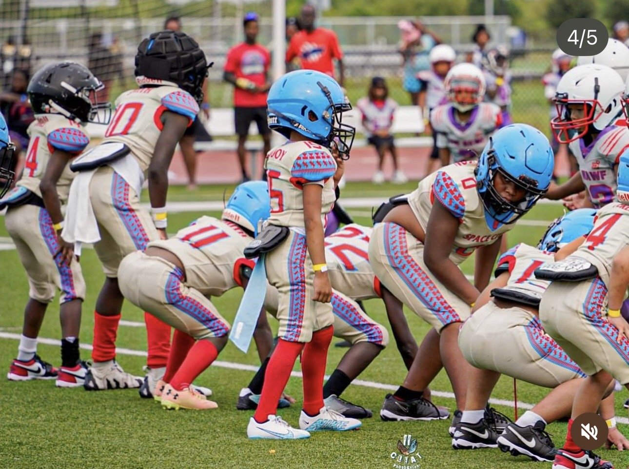 Young football players in beige uniforms with blue and red accents, wearing blue helmets, are lined up on a football field during a game. Some players are in stance, ready for the next play, while others are standing or crouching. Spectators are visi