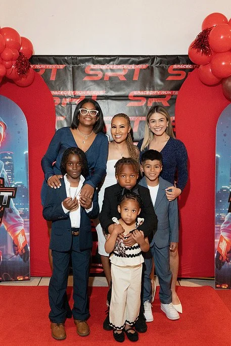 Group of six people, including children and adults, standing together on a red carpet in front of a SpaceX SRT backdrop decorated with red balloons.