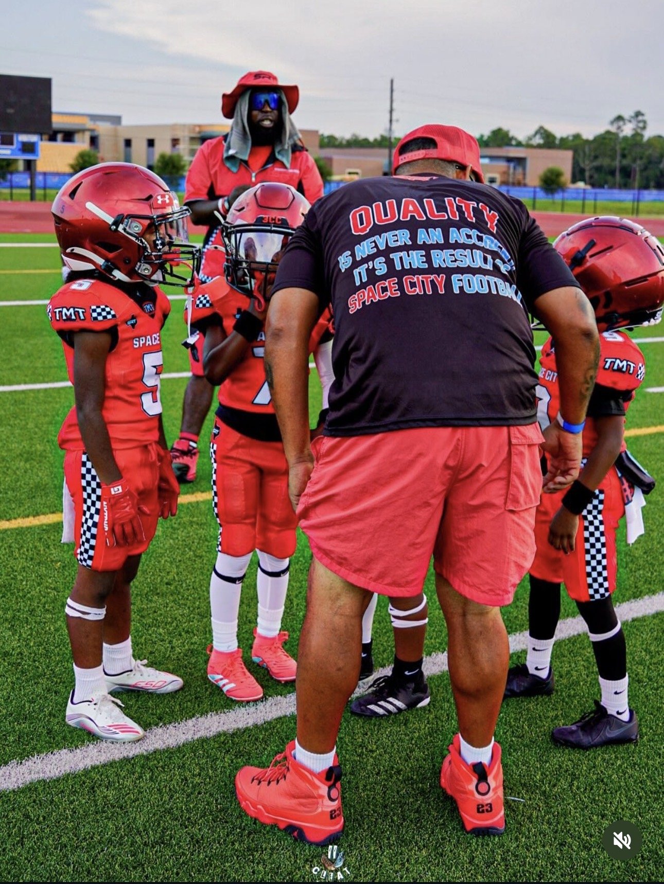 A youth football team in red uniforms standing on a football field, listening to their coach who is giving instructions during a game or practice.