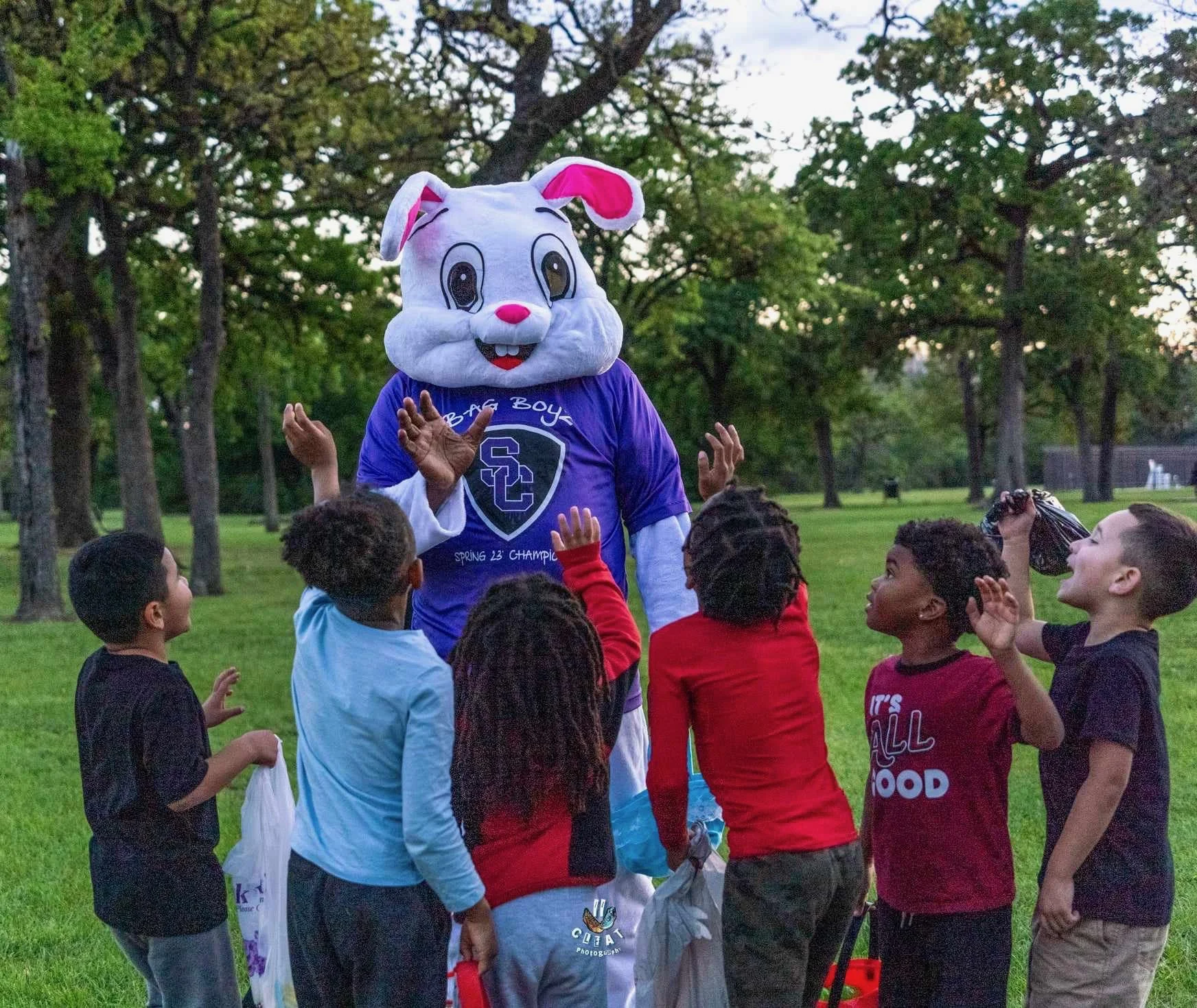 A person in a large white bunny costume with pink accents and a purple T-shirt, surrounded by children outdoors in a park, engaging in an activity.