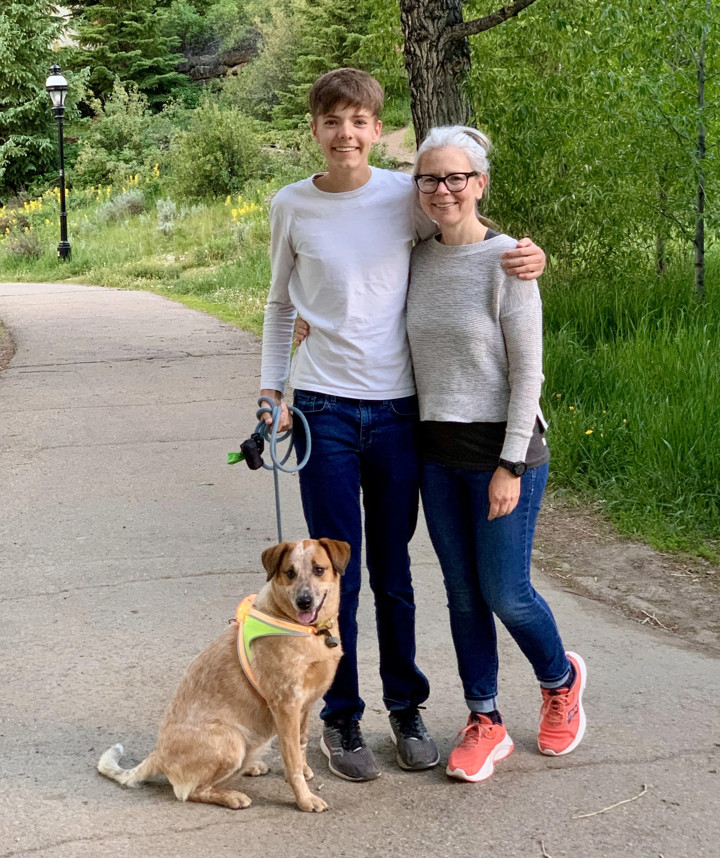 A young man and an older woman smiling and embracing each other outdoors on a walking path in a park, with a dog sitting on the ground in front of them. The young man is wearing a white long-sleeve shirt, jeans, and sneakers, and the woman is wearing glasses, a gray sweater, jeans, and sneakers. The dog is a small, tan-colored mixed breed wearing a neon yellow harness. Green trees and bushes surround them.