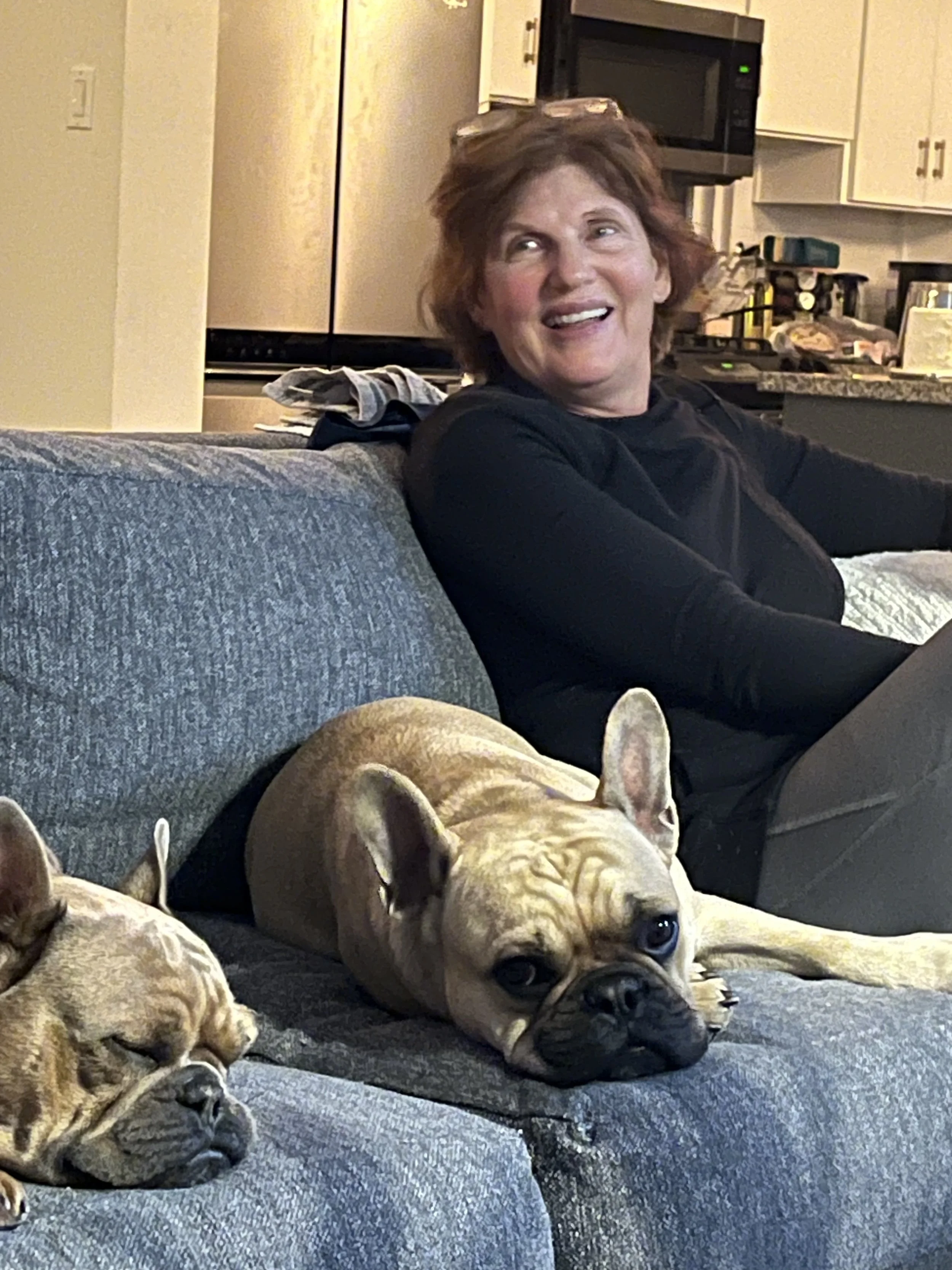 A woman with short red hair sitting on a blue couch, smiling and looking to her left, with two French bulldogs lying on the couch in front of her, one sleeping and the other looking at the camera.