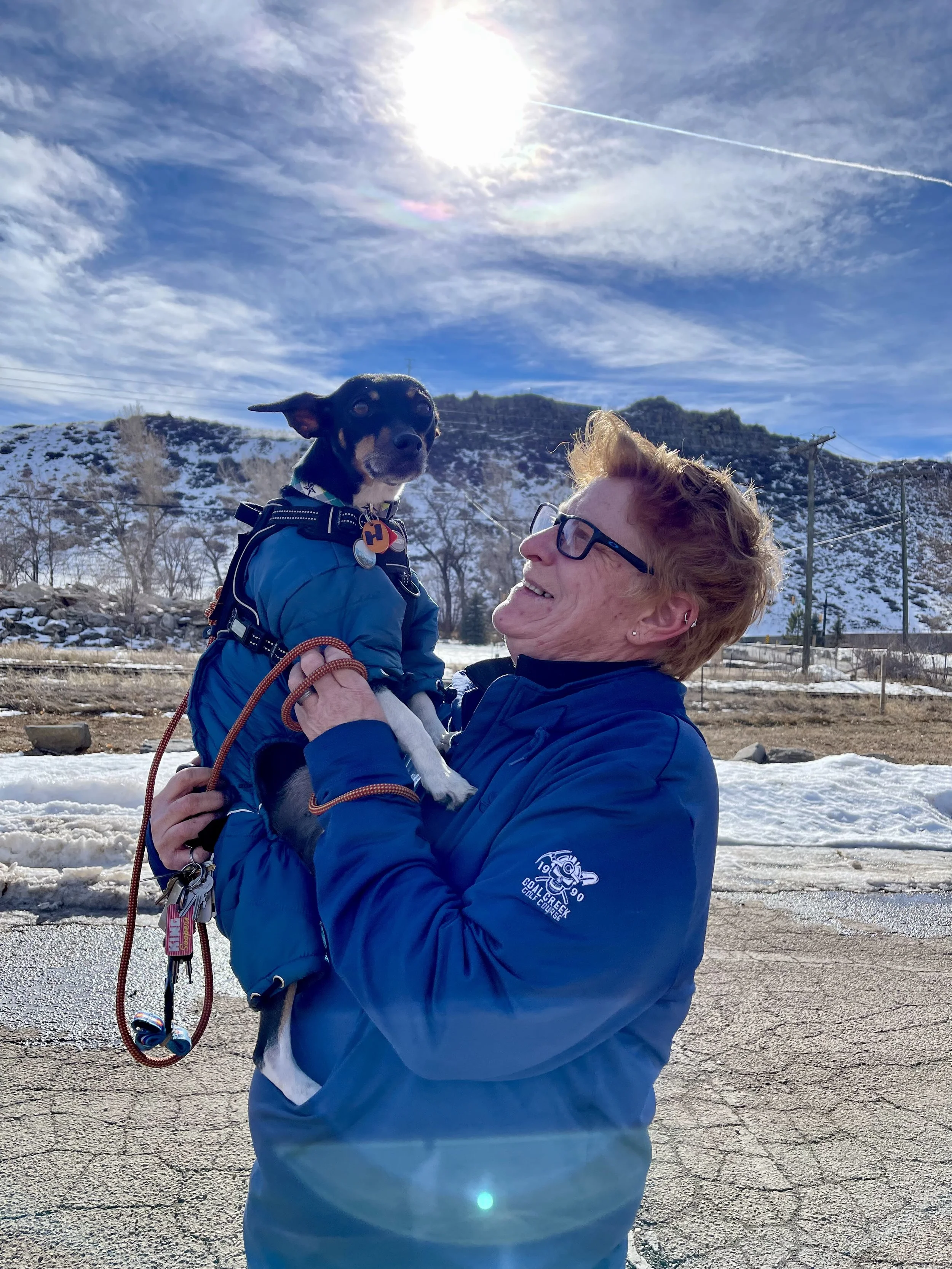 A woman wearing a blue jacket and glasses holds a small black and tan dog wearing a blue jacket, outdoors on a sunny winter day with snow on the ground and hills in the background.
