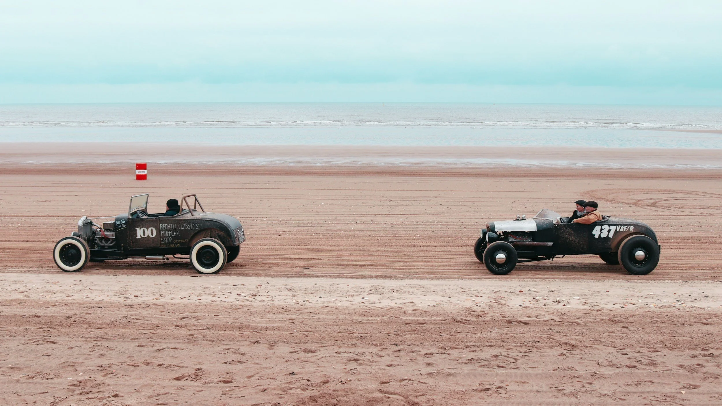 Two vintage cars racing on a sandy beach, with the ocean in the background.