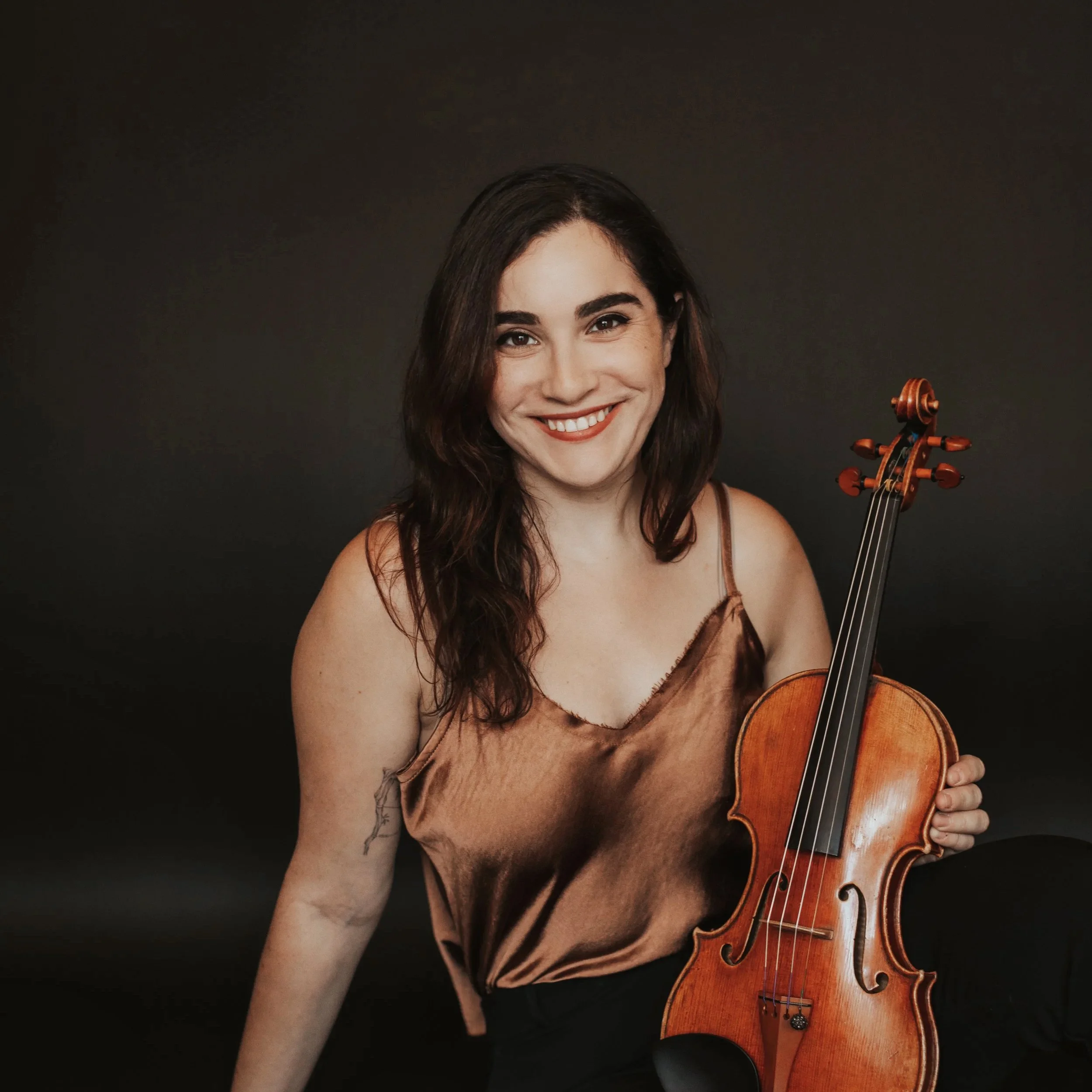A woman with dark wavy hair holding a violin and smiling, wearing a satin brown top, against a dark background.