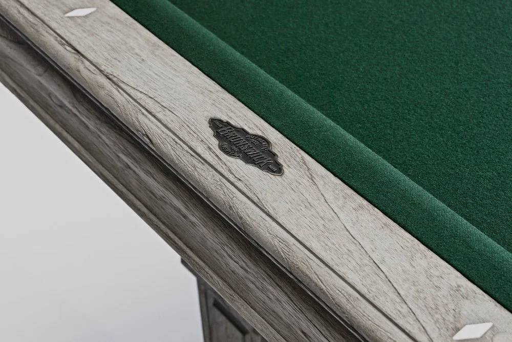 Close-up of a vintage billiards or pool table with a green felt surface and weathered whitewashed wooden edge, featuring a small metallic emblem.