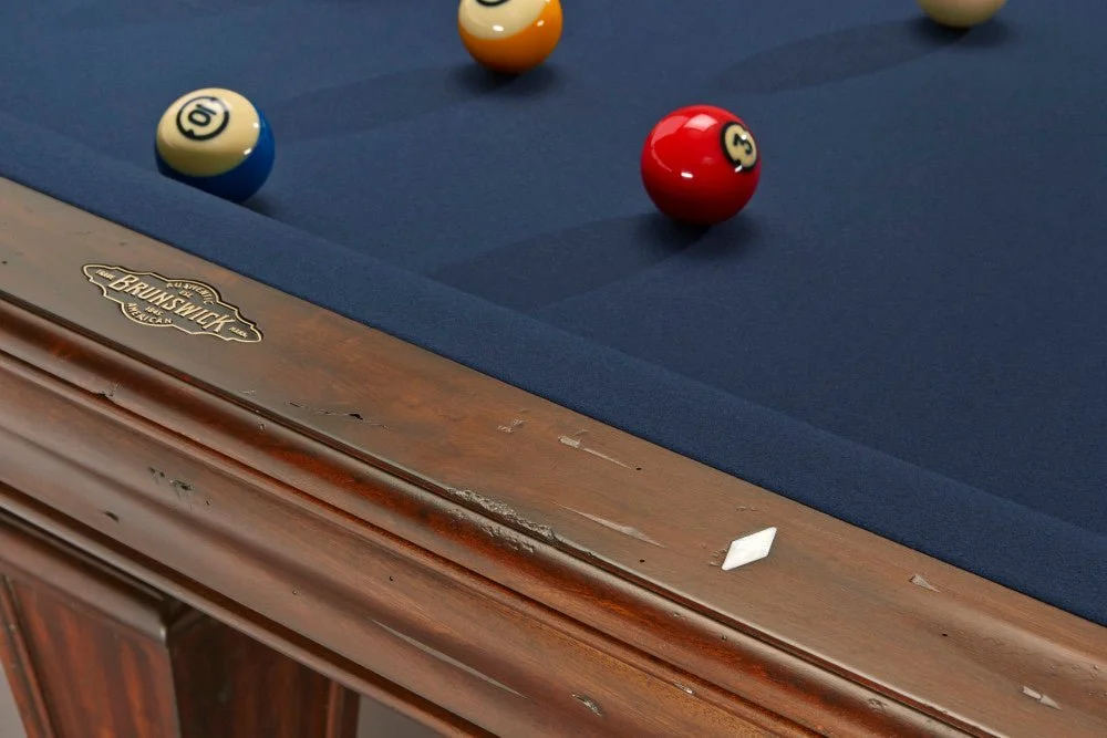 Close-up of a pool table with a blue felt surface and white, red, and yellow balls scattered on it, and part of the wooden frame with a 'Brunswick' label.