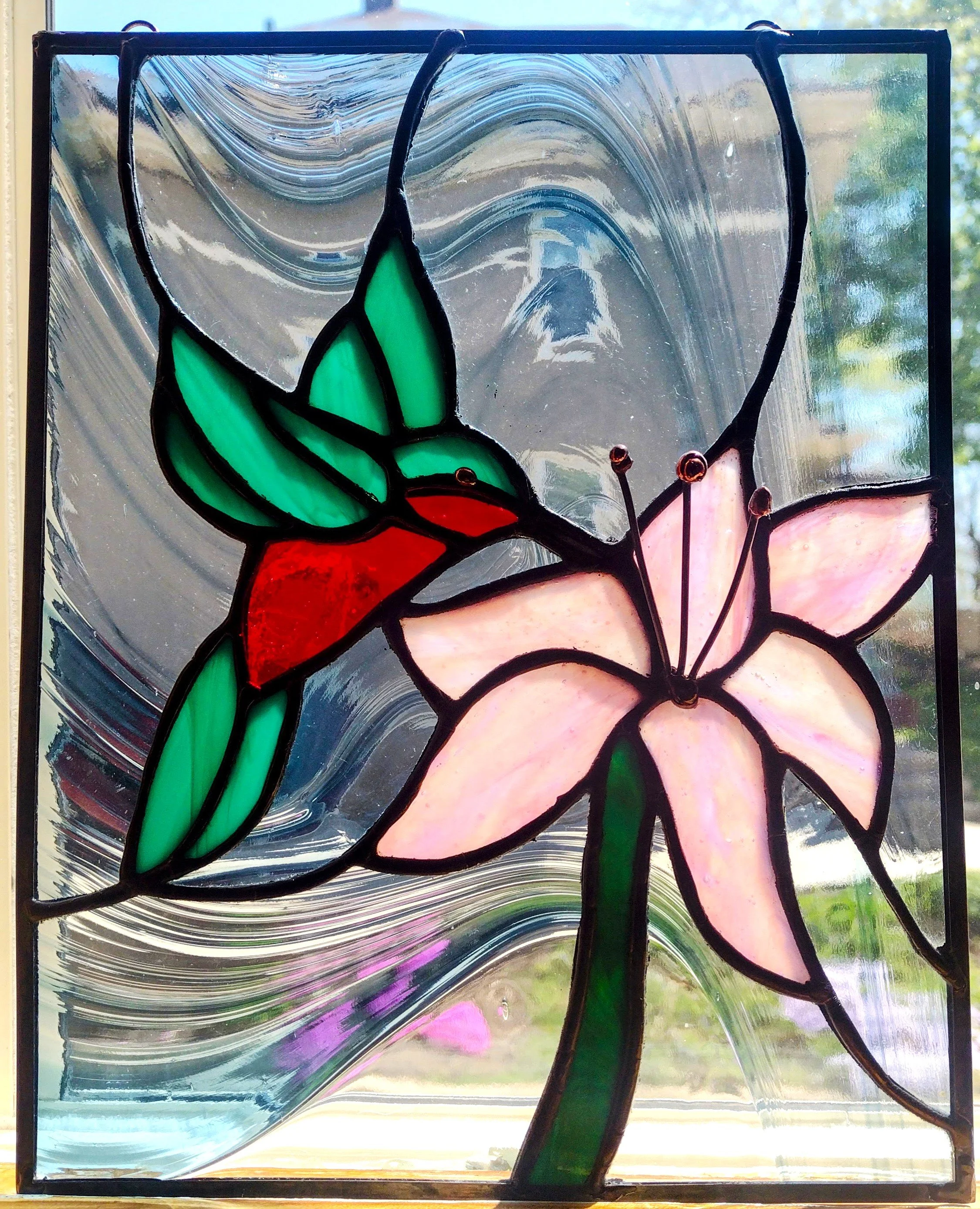 Colorful stained glass window featuring a pink lily flower with green leaves and a red hummingbird on a glass background.
