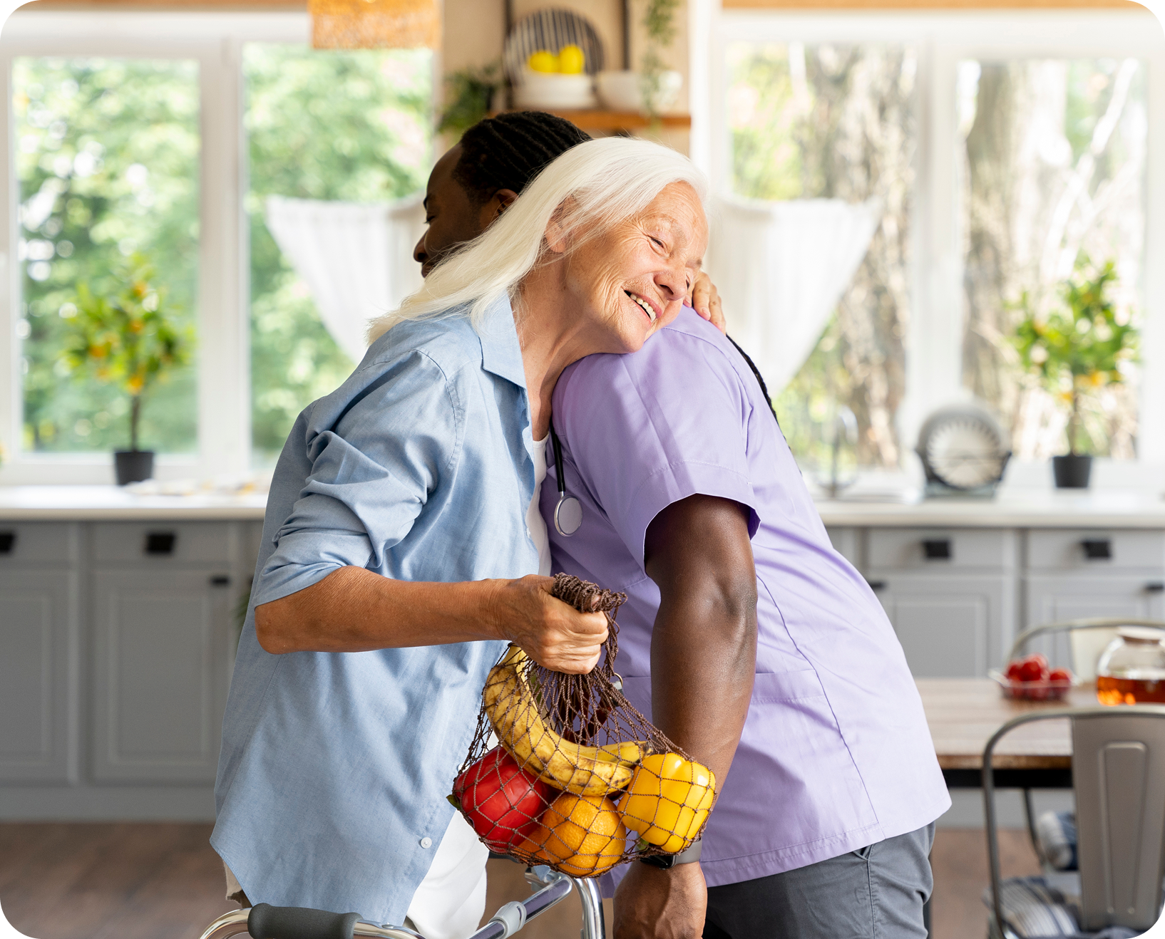 An elderly woman in a light blue shirt smiling and hugging a healthcare worker with a purple scrubs. The elderly woman holds a shopping bag with bananas, apples, and oranges, in a bright kitchen with large windows and green plants.