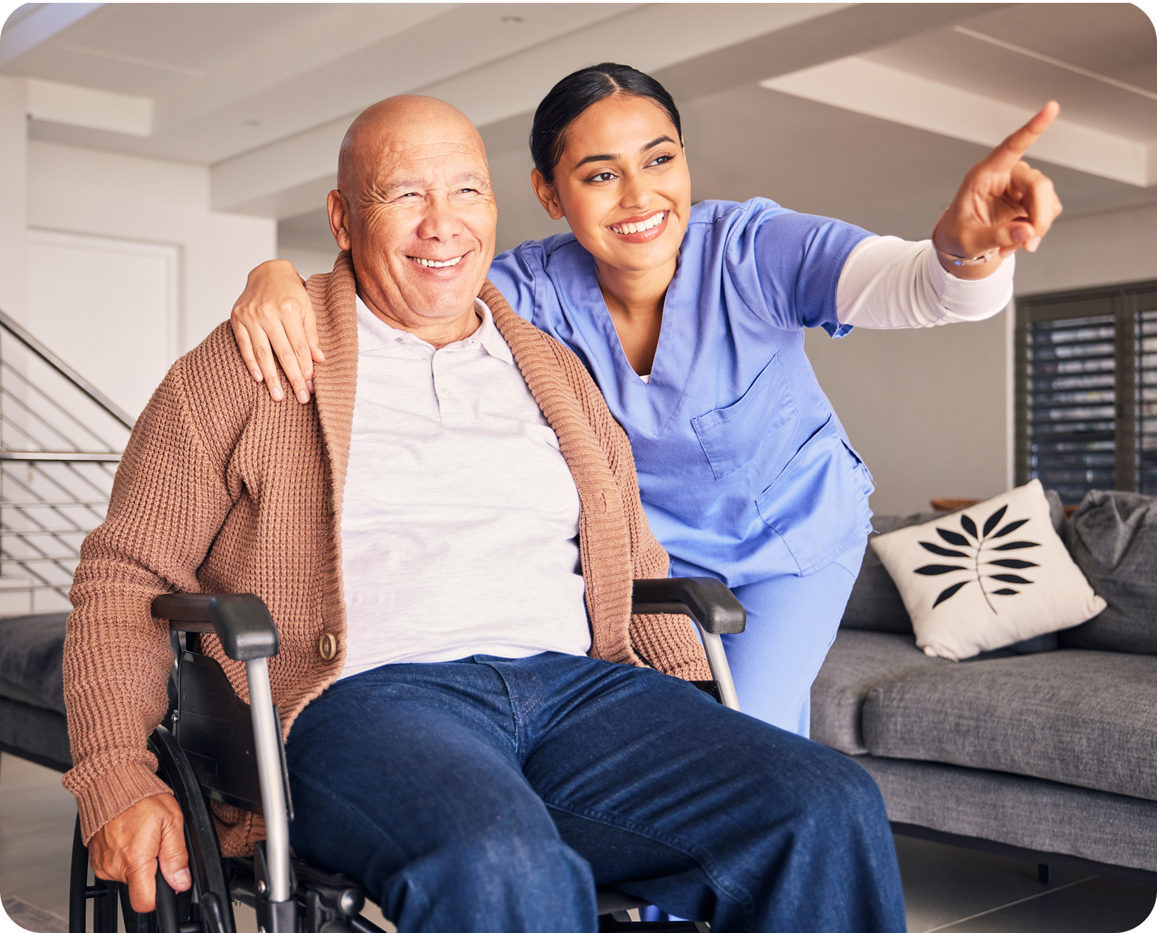 A nurse in blue scrubs smiling and pointing, standing beside a smiling elderly man in a wheelchair with a patterned pillow on a gray sofa in a modern living room.