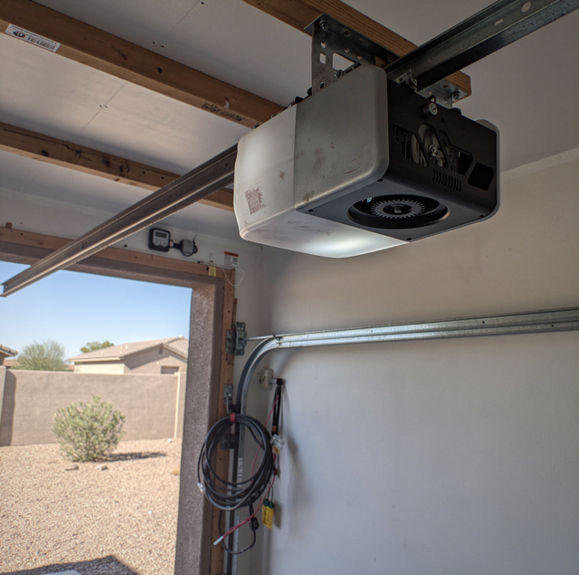 Garage door opener mounted to ceiling with metal rail and black motor unit above white drywall walls.