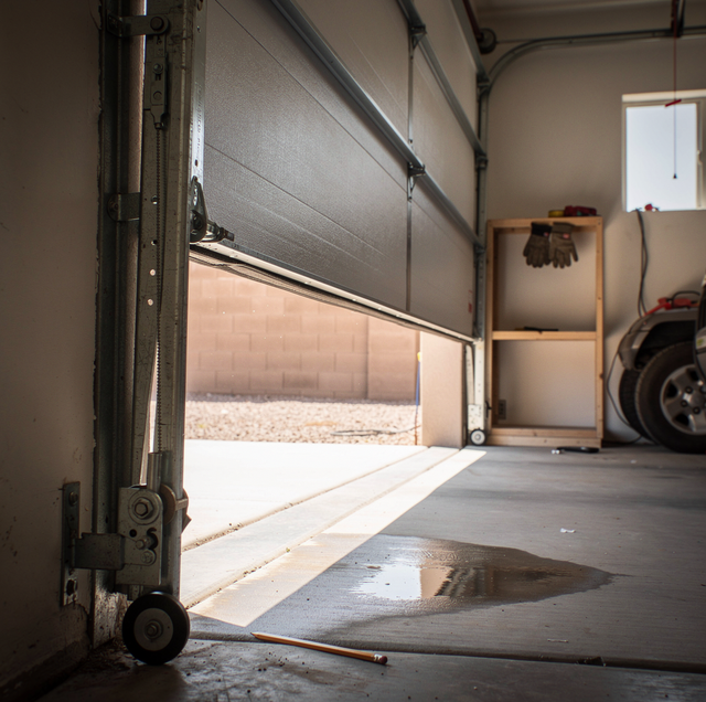 Open garage door with shelving unit, gloves, tire, and puddle on concrete floor inside