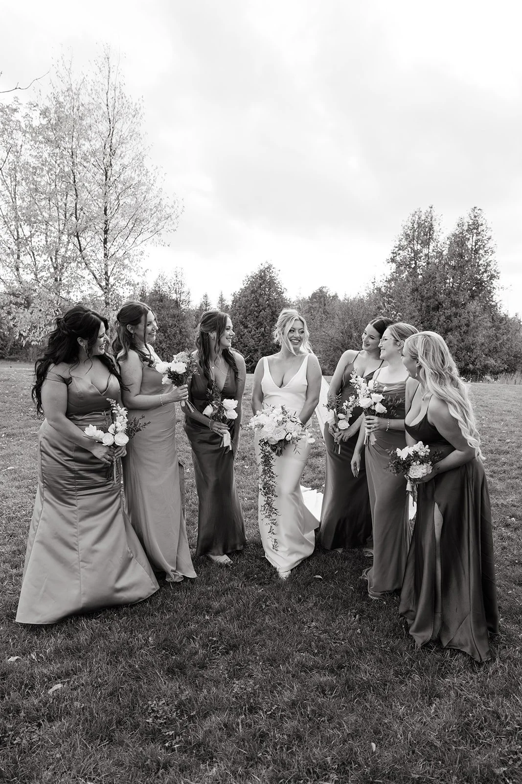 A bride and seven bridesmaids standing outdoors on grass, holding bouquets, smiling and talking. The photo is black and white, with trees and cloudy sky in the background.