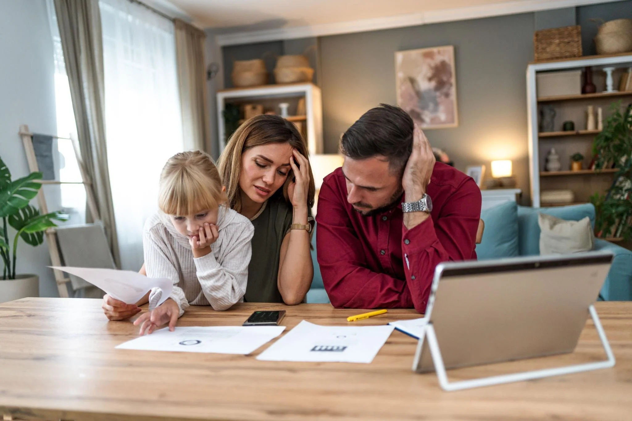A man and woman looking stressed while reviewing financial paperwork and bills at a wooden table with a young child.