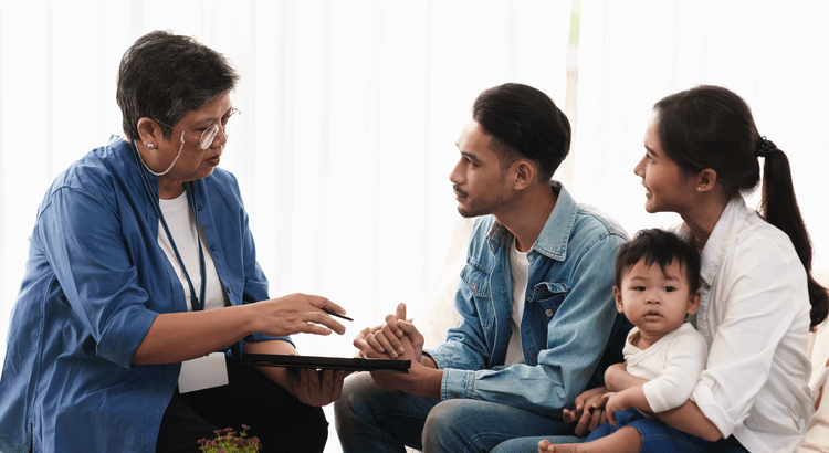 A young couple with a toddler sitting on a couch and listening to a professional real estate agent or financial advisor explain home buying documents on a tablet.