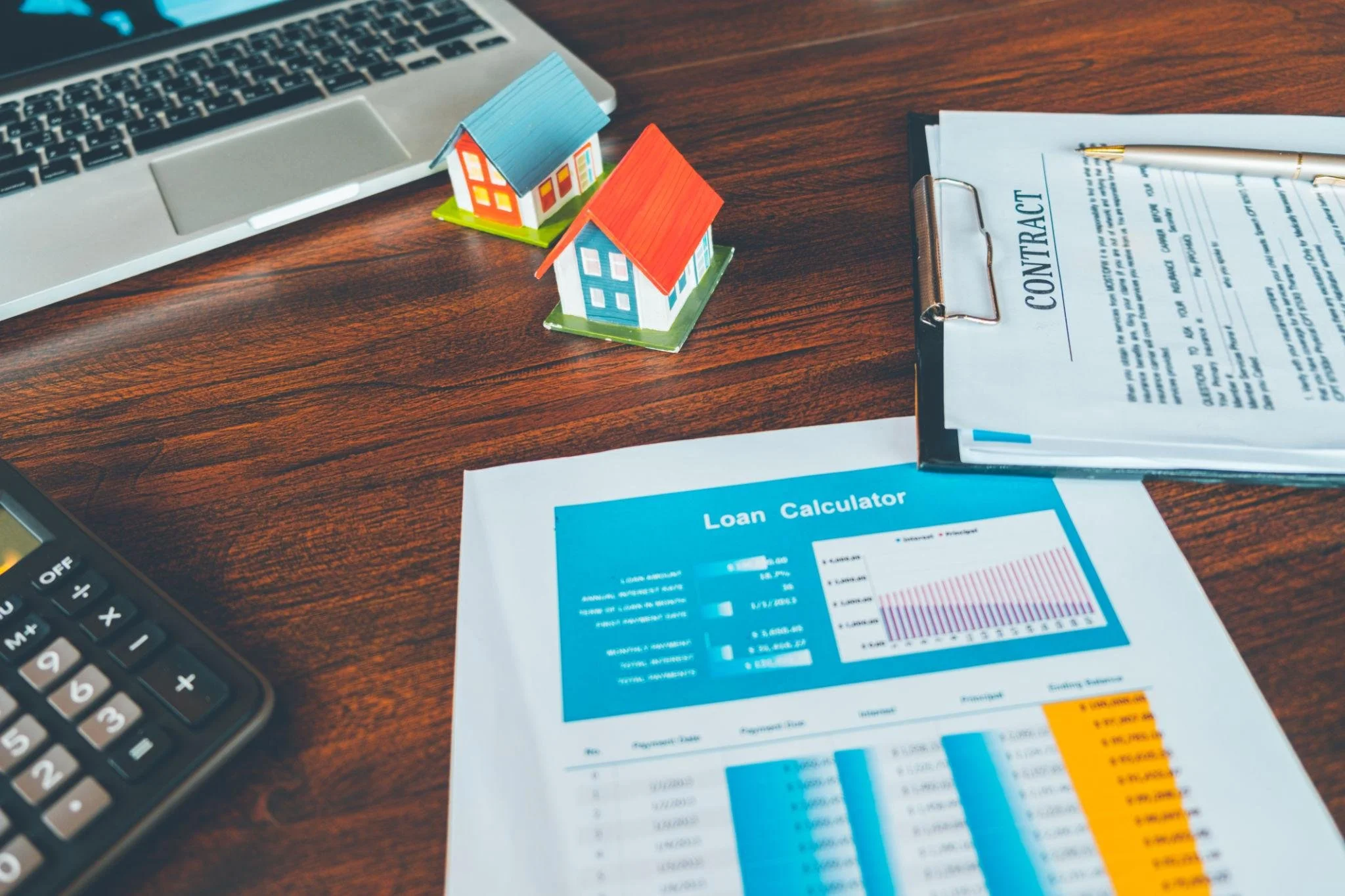 A desk with two small toy houses, a formal real estate contract, a calculator, and a printed mortgage loan calculator sheet showing monthly payment breakdowns.