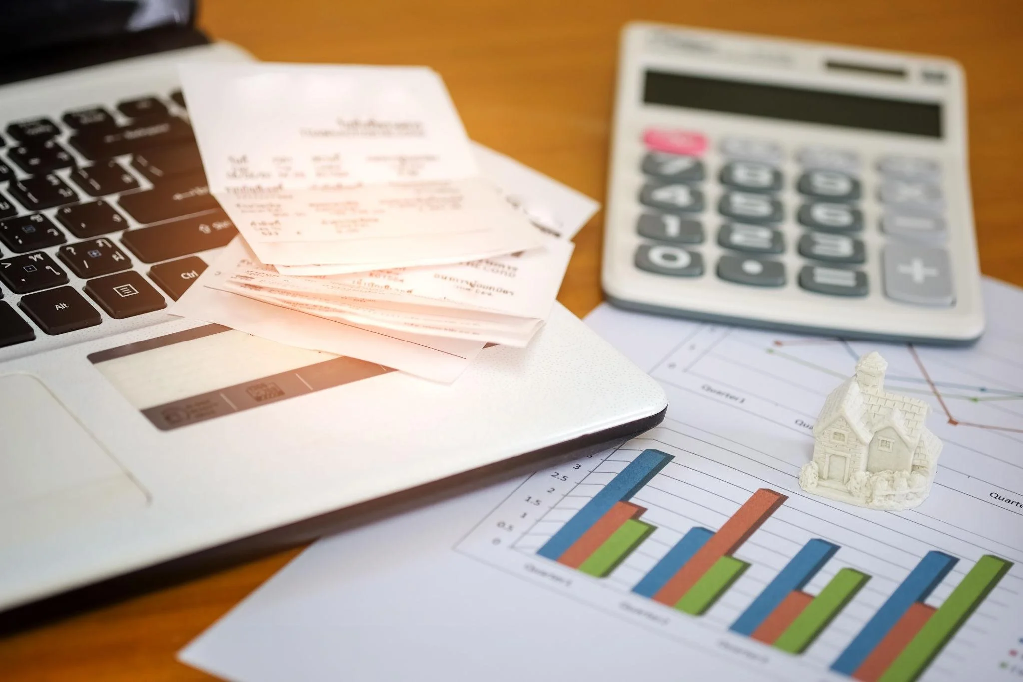 A laptop keyboard next to a stack of receipts, a white calculator, and a bar graph showing financial growth with a small house model.