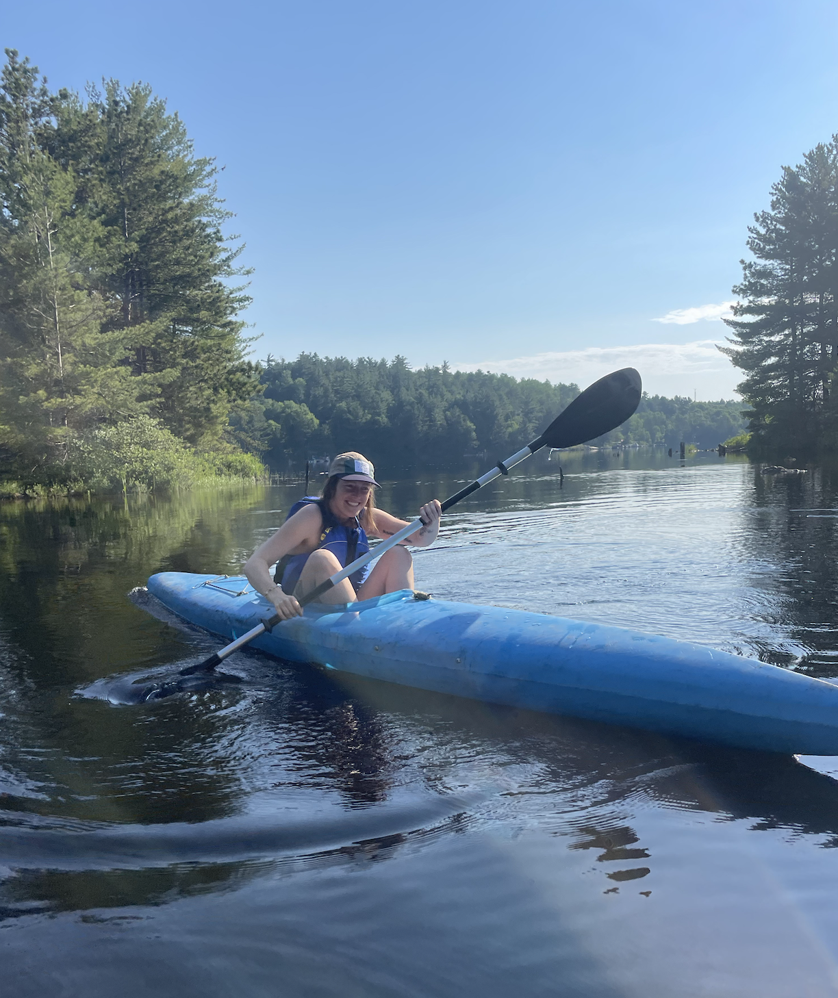 A woman in a blue kayak paddling on a calm river with green trees and a blue sky in the background.