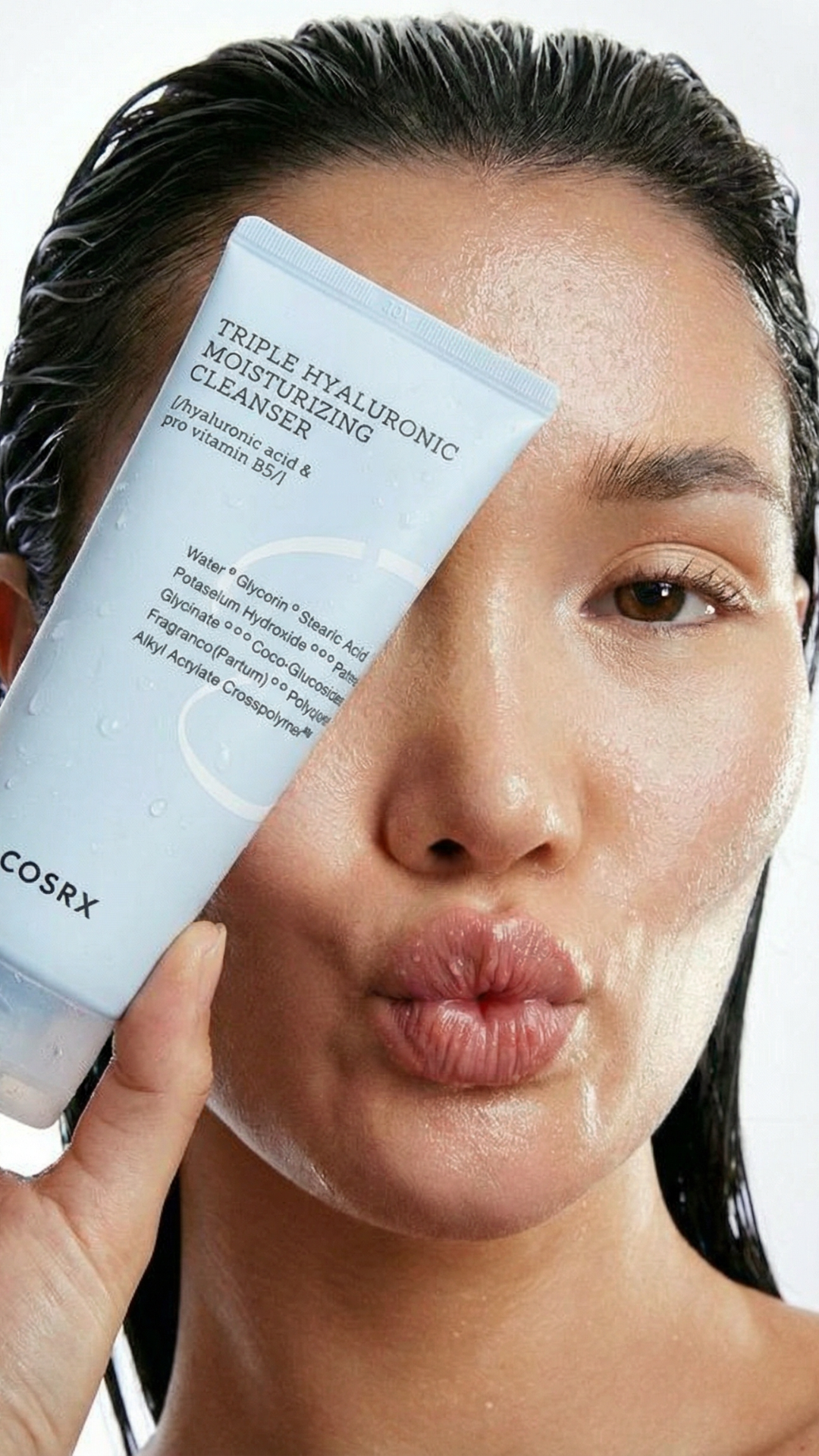 A woman with wet hair holding a skincare product near her face, making a kissing face.