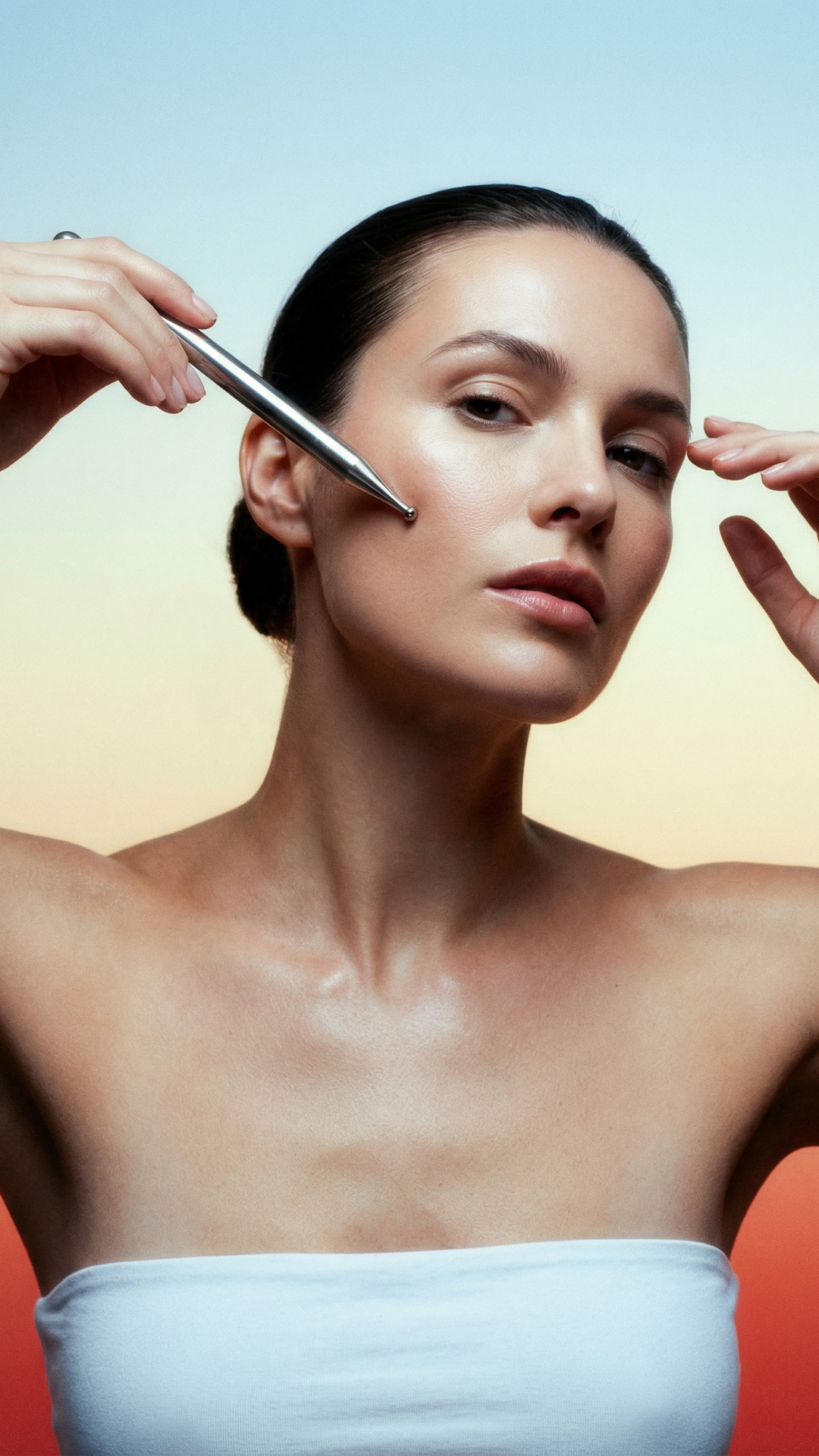 A woman with dark hair pulled back, applying skin treatment with a silver tool to her cheek, against a pastel-colored background.