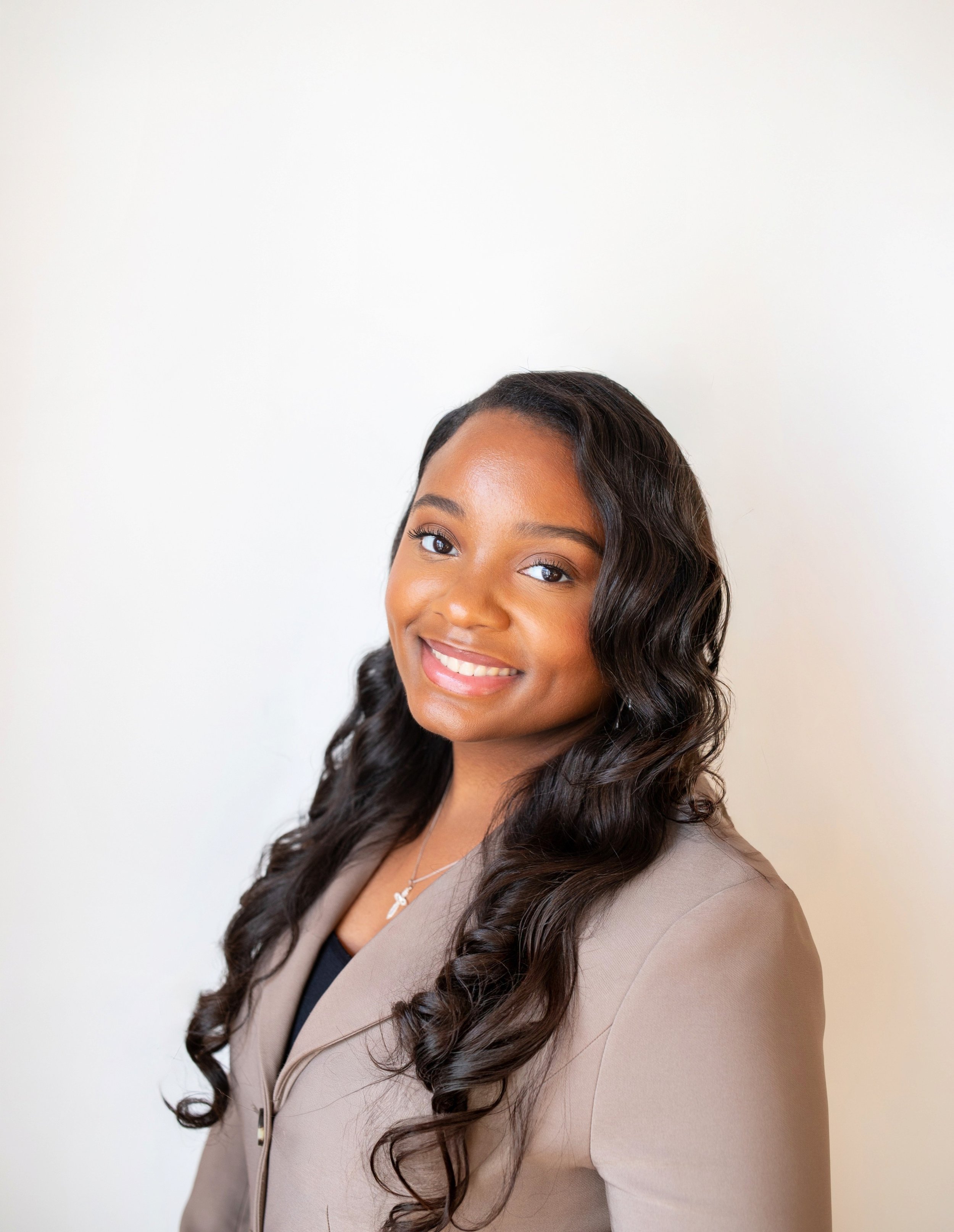 Close-up of a smiling woman with wavy black hair, wearing a beige blazer over a black top, and a silver necklace with a small charm, standing against a plain white background.
