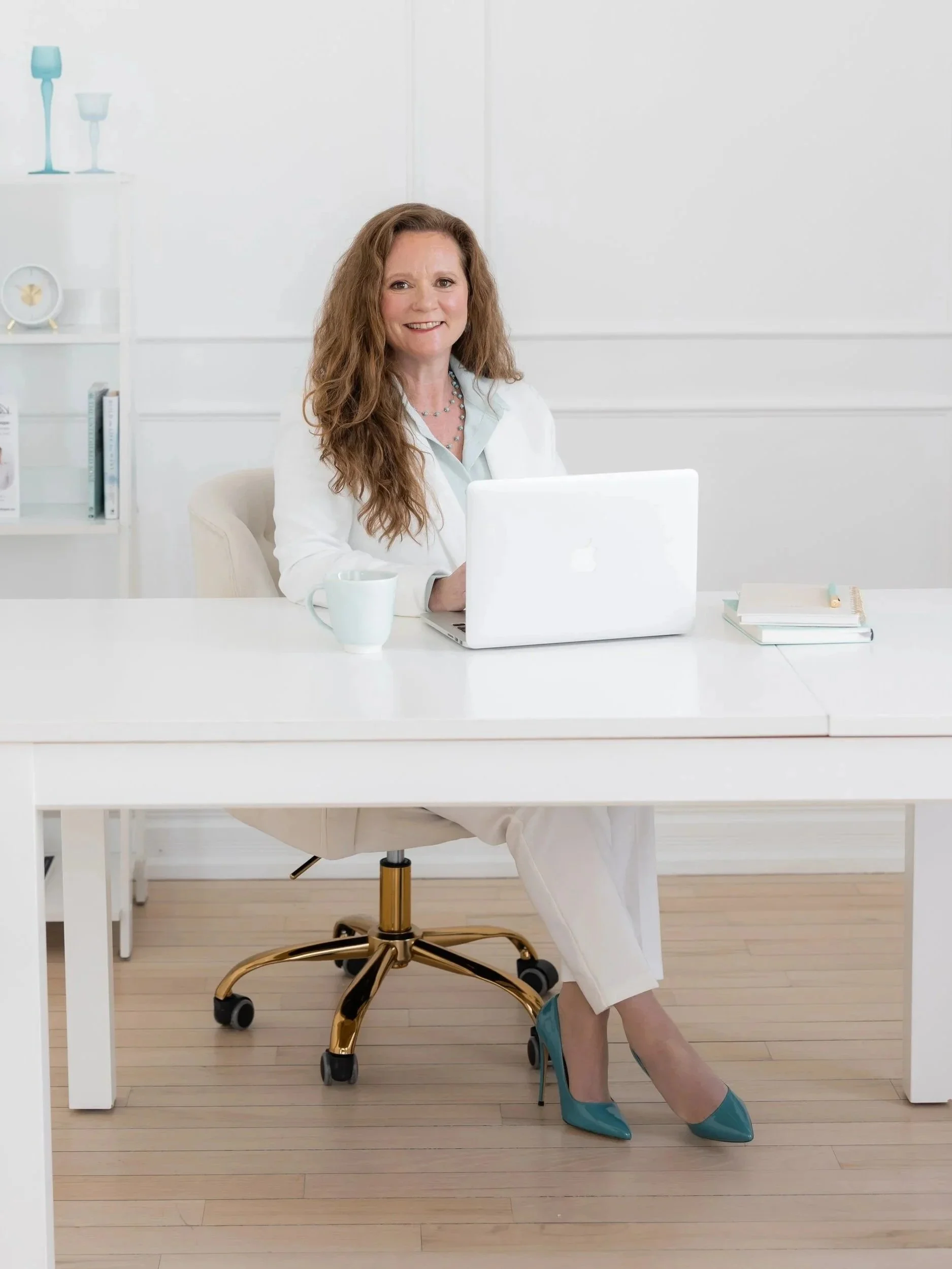 A woman with curly brown hair seated at a white office desk wearing teal high heels and a white blazer. She has a smile and is working on a silver laptop. There is a white coffee mug and a closed notebook on the desk. The background features white shelves with decorative items and books, and a white wall with paneling.