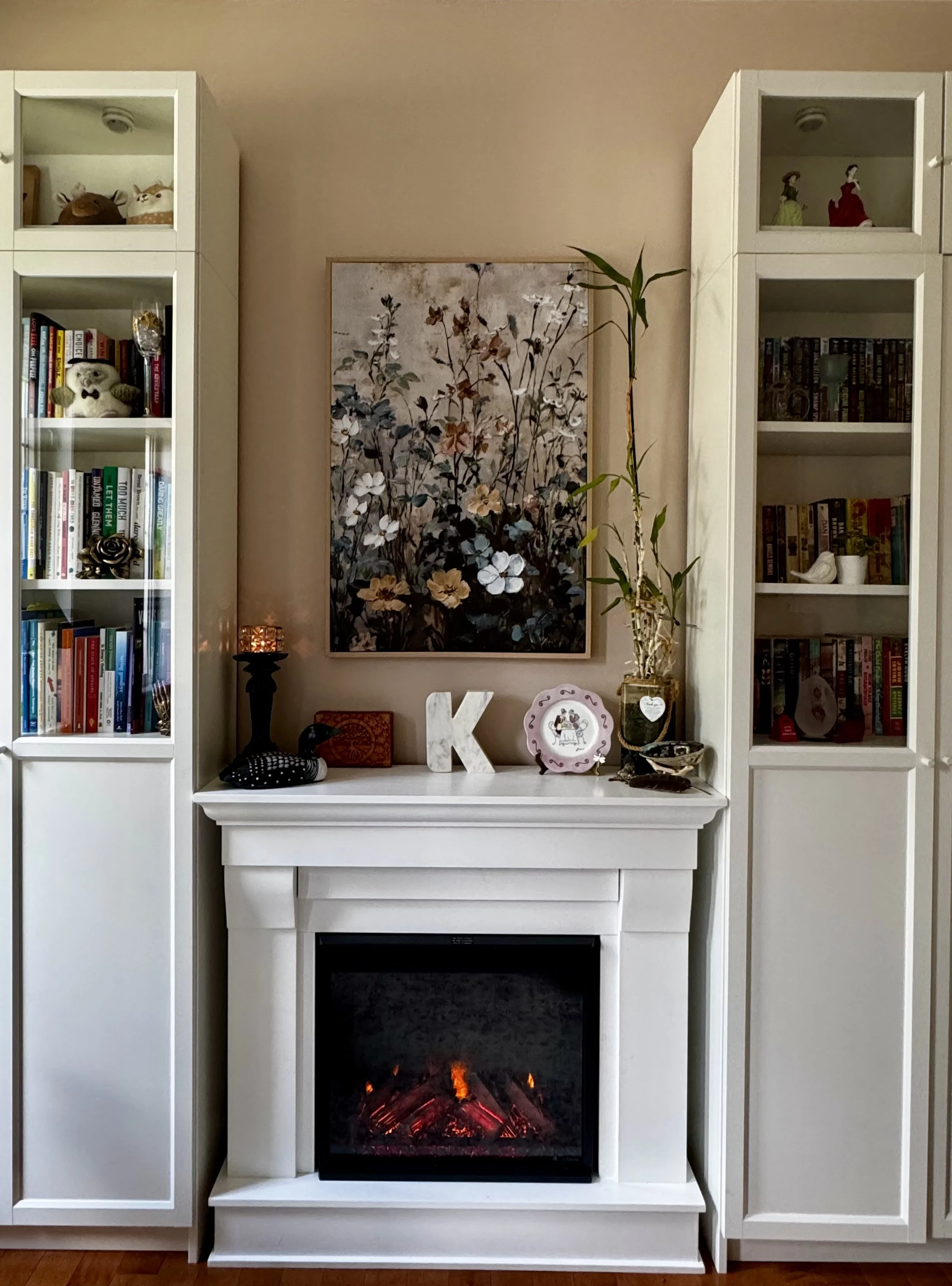 White entertainment center with a fireplace in the middle, decorated with books, plush toys, and decorative items, with two tall cabinets on either side containing figurines, books, and a ceramic bird.