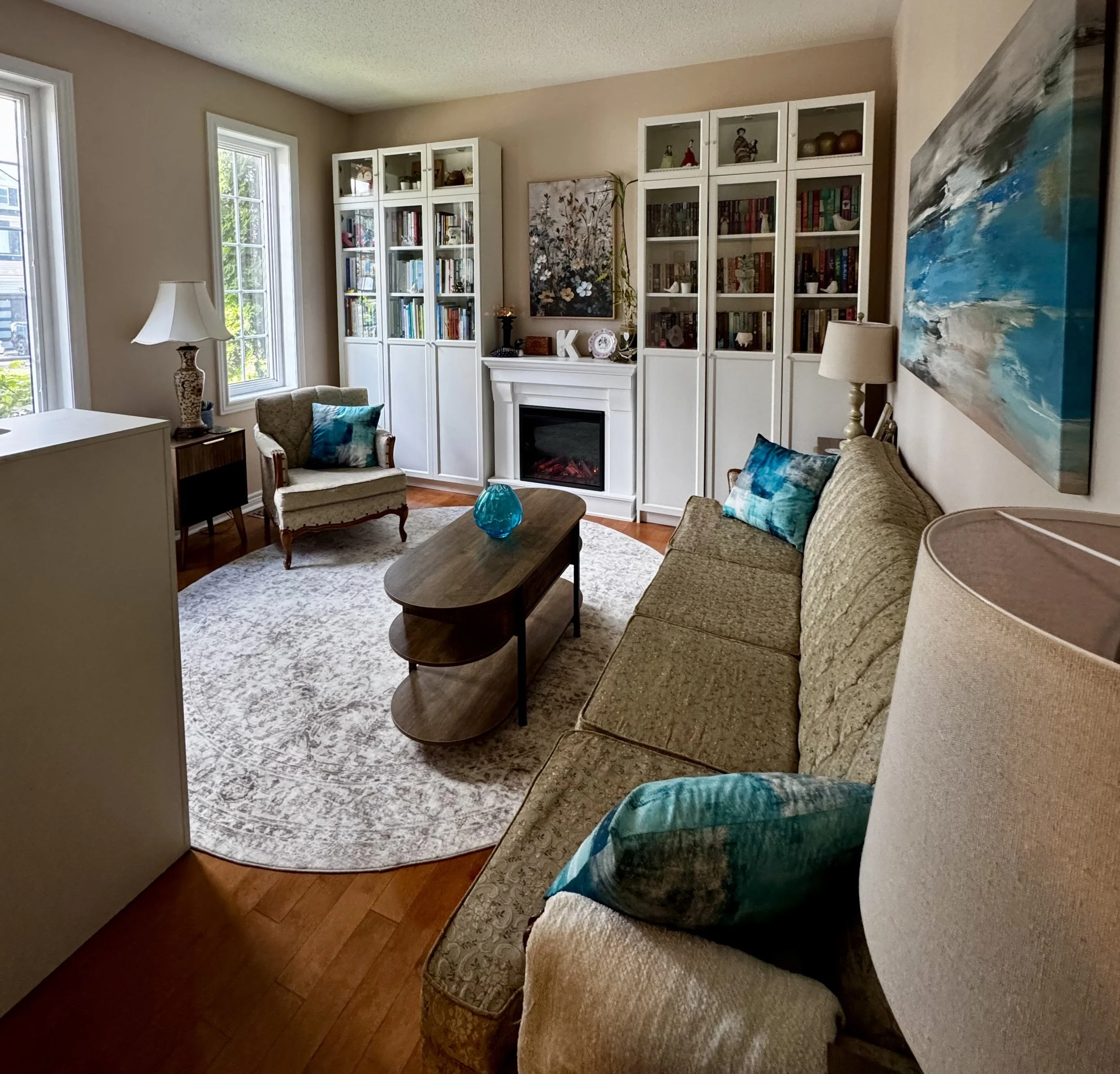 A cozy living room with beige walls, two large windows, a beige sofa with blue pillows, an armchair with a patterned cushion, a wooden coffee table with a blue glass vase, white built-in bookshelves filled with books and decor, a white fireplace, and abstract wall art.