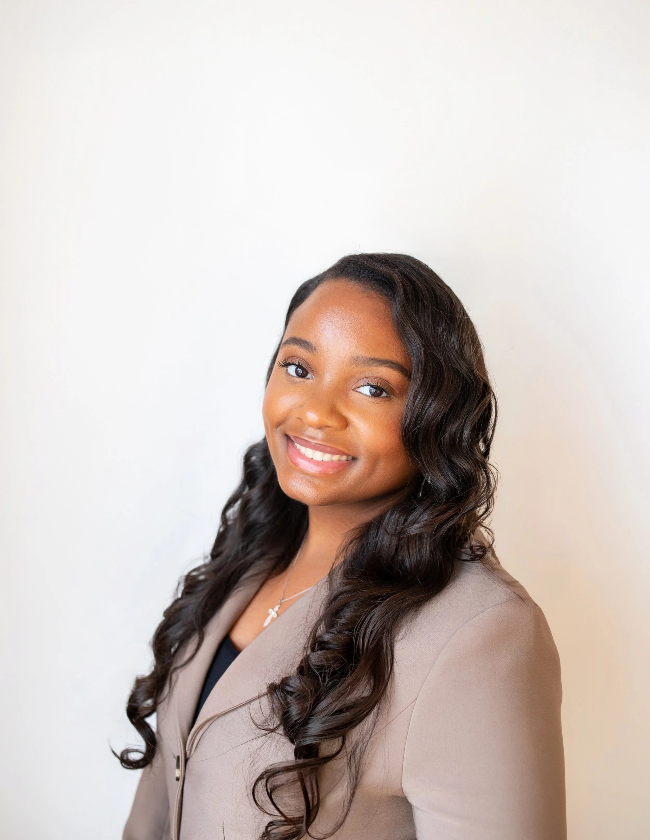 A young woman with long, dark, wavy hair smiles at the camera. She is wearing a beige blazer over a black top, and a delicate necklace with a small cross pendant. The background is plain white.