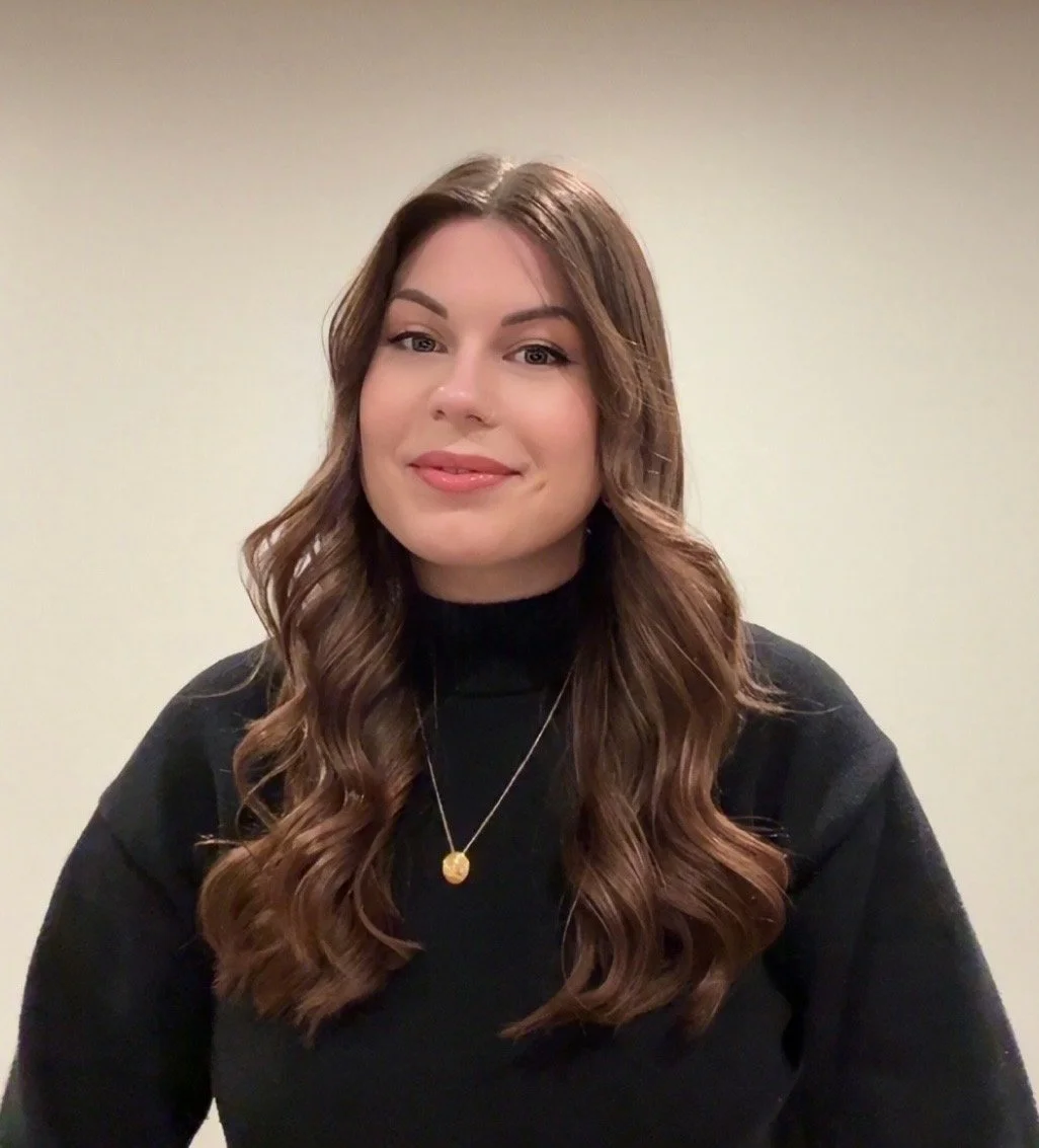 A woman with long, wavy brown hair, wearing a black sweater and a gold pendant necklace, smiling in front of a plain beige wall.