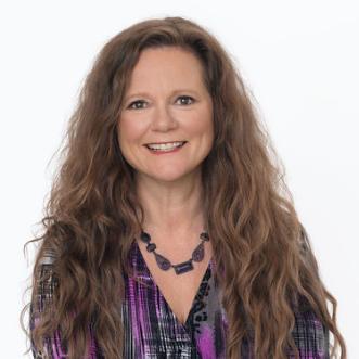 A woman with long, curly brown hair, smiling, wearing a purple and black patterned shirt and a purple beaded necklace.