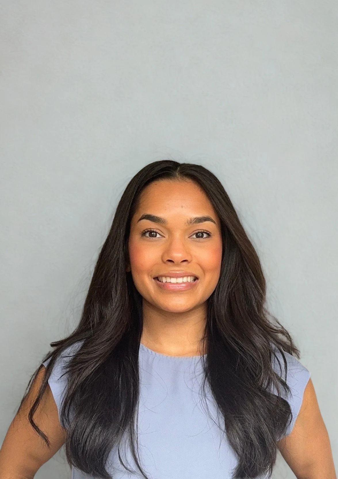 A woman with long dark hair and a smile, wearing a light blue sleeveless top, standing in front of a plain light gray wall.