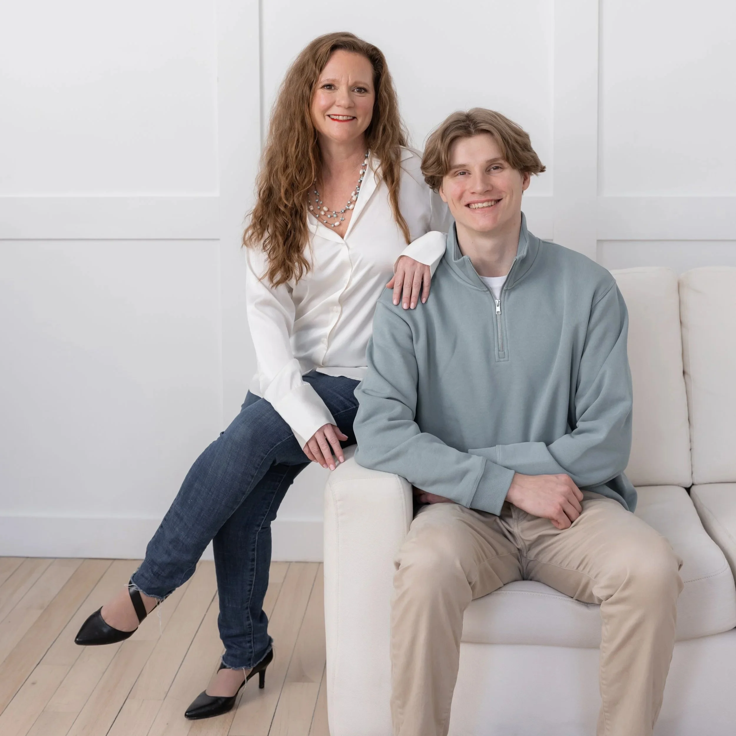 A woman with long wavy hair and a necklace, wearing a white blouse, standing with one leg crossed, smiling. Next to her, a young man with wavy hair, wearing a gray hoodie and beige pants, sitting on a white chair, smiling. The background is white with paneled walls, and the floor is wooden.