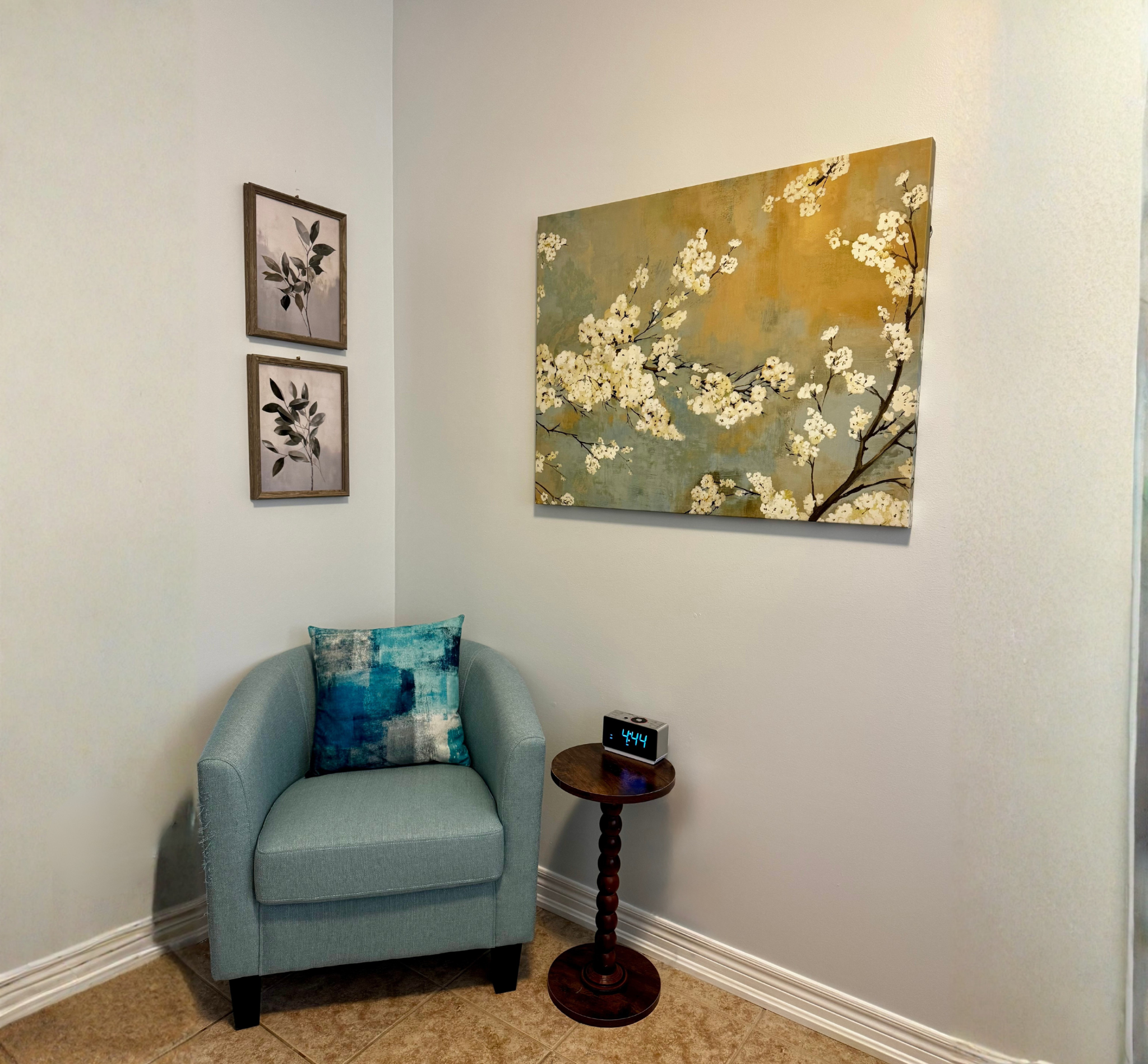 A cozy corner with a teal upholstered armchair with a patchwork blue, white, and gray cushion, a wooden side table with a digital clock showing 4:44, a wall with three framed botanical prints, and a large painting of white cherry blossoms on a muted background.