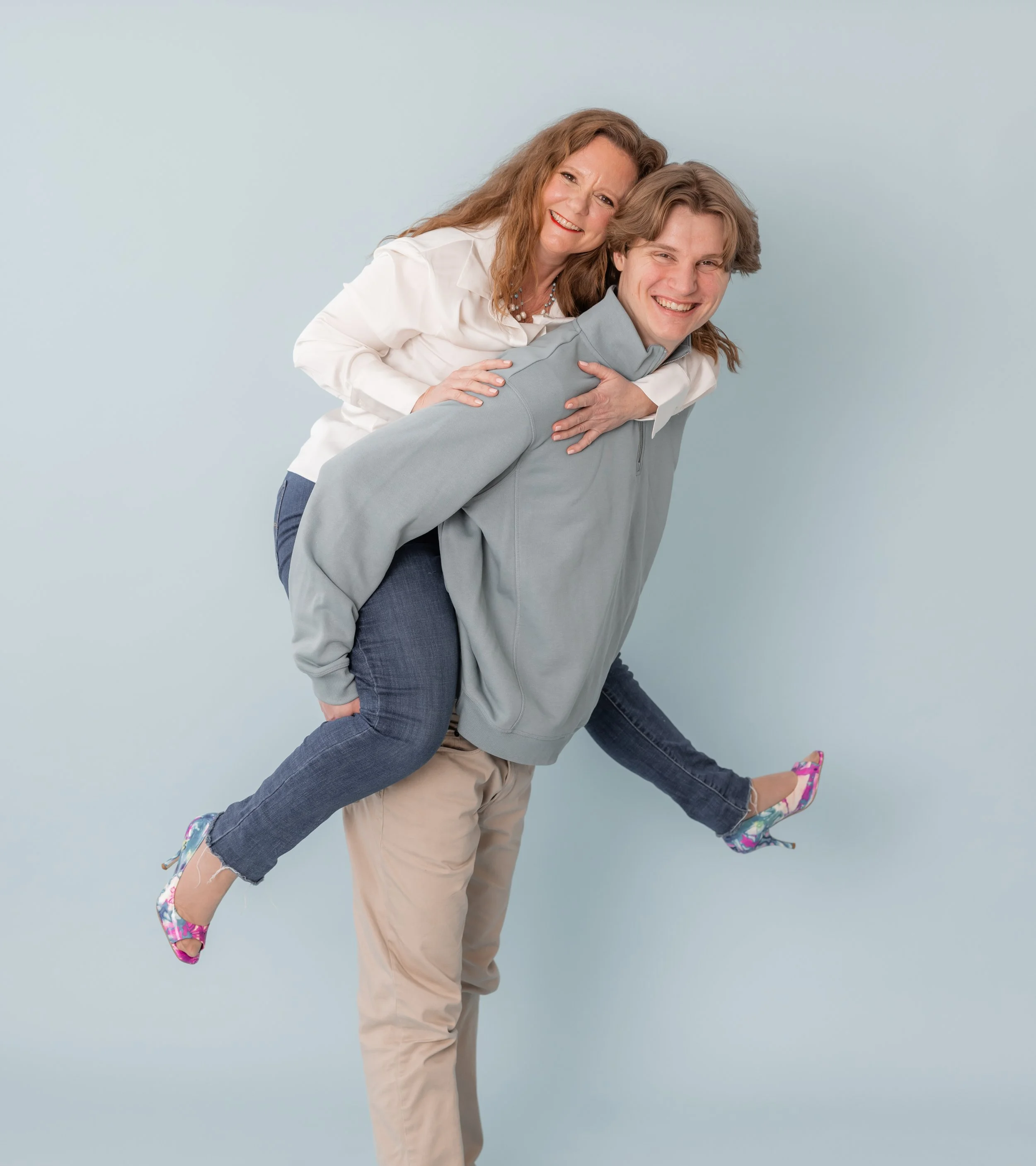A woman giving a piggyback ride to a young man, both smiling and laughing against a light blue background.