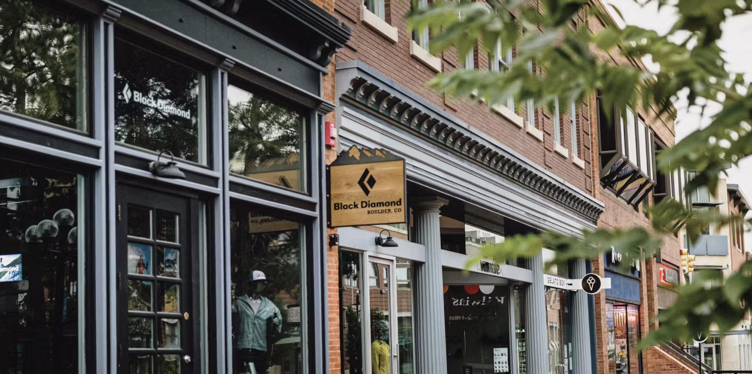 Exterior of a retail building with glass windows, brick walls, and awnings. Signs for Black Diamond Boulder, CO, and other stores are visible. Green trees partially obscure the view.
