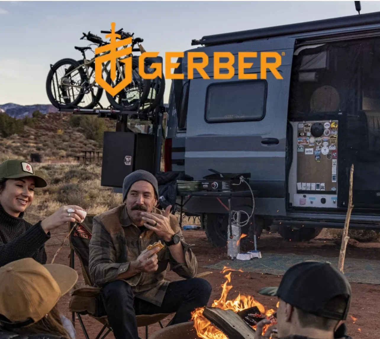 People sitting around a campfire near a camper van with a bike rack, in a desert landscape during sunset.