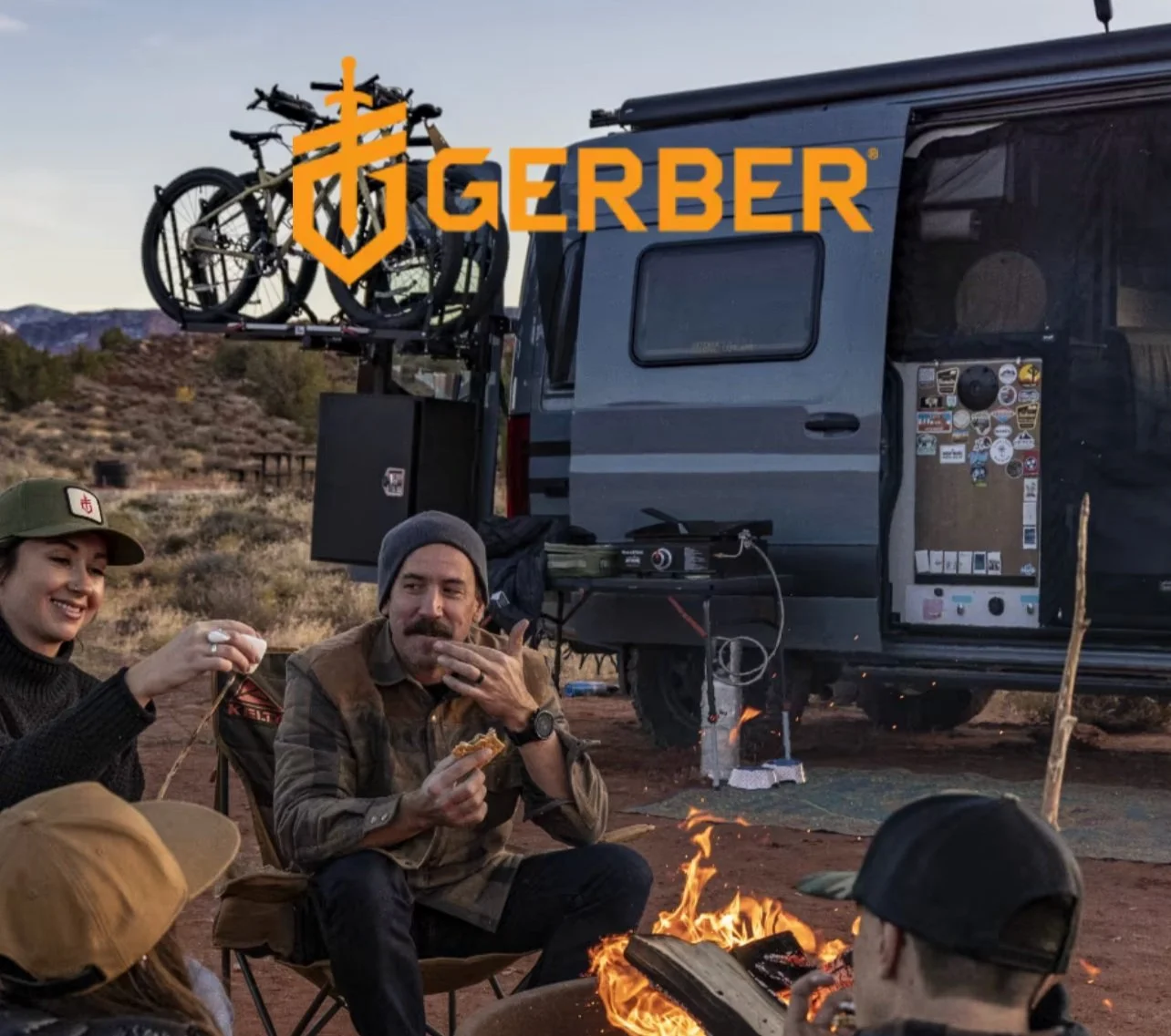 People camping outdoors near a van with bicycles on a rack, sitting around a campfire, in a desert landscape during sunset.
