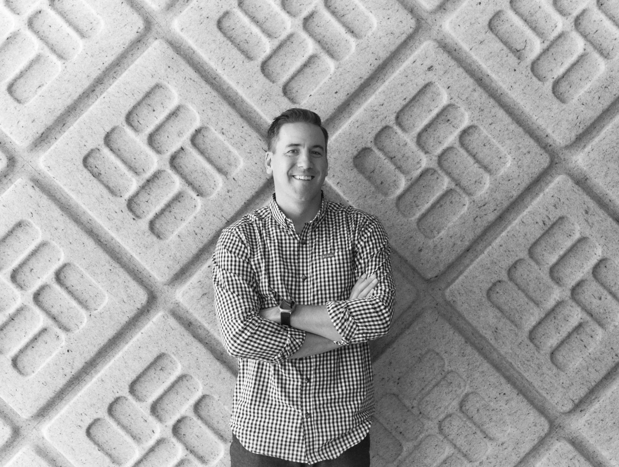 A young man wearing a checkered shirt with sleeves rolled up, smiling and crossing his arms, standing in front of a textured wall with geometric patterns.